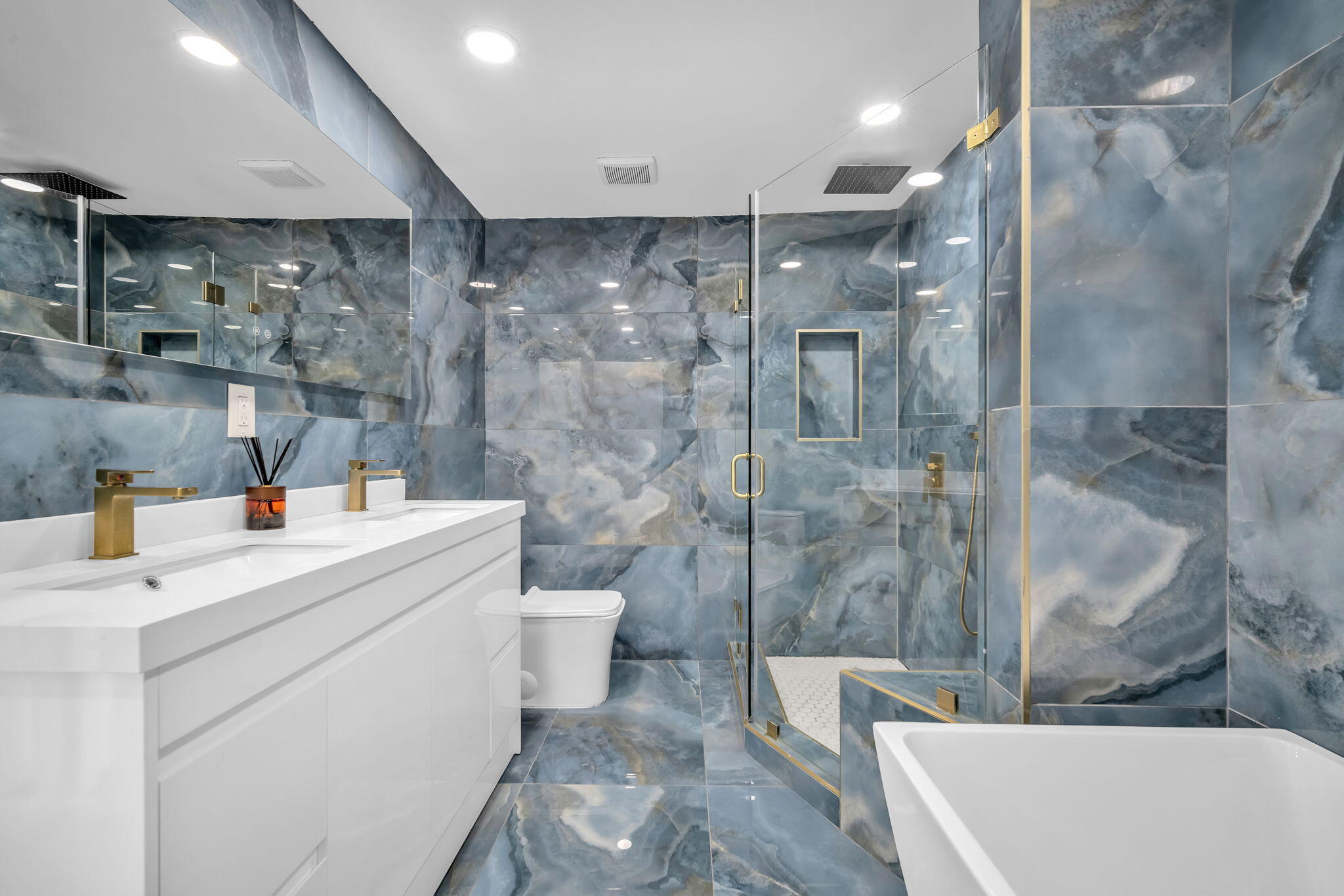 Bathroom featuring full-height blue decorative stone tiling, white double-vanity cabinet with integrated sinks, brass-finish fixtures, glass-enclosed walk-in shower with built-in recessed shelving, and a white freestanding soaking tub