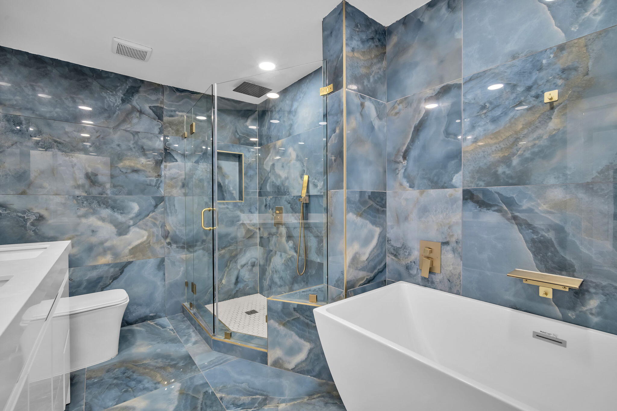 Large-format blue stone wall and floor tiling, freestanding white soaking tub, walk-in glass-enclosed shower with a recessed niche, and gold-finish plumbing fixtures