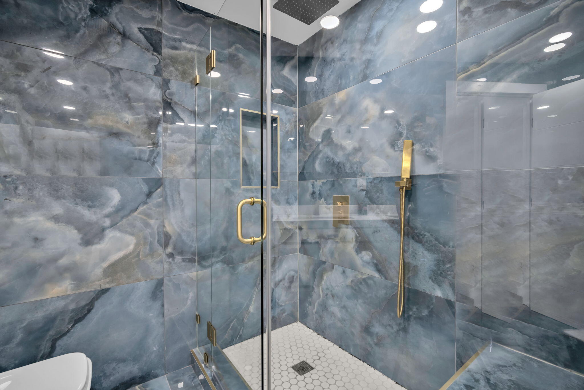 Walk-in shower featuring large-format blue stone-finish wall tile, gold-toned hardware, rainfall showerhead, integrated wall niche, and hexagonal floor tile