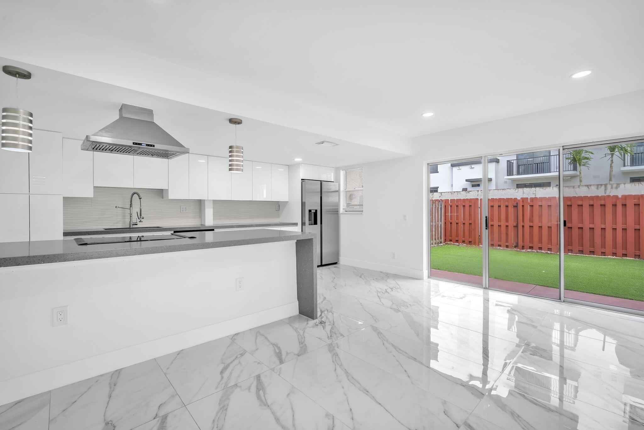 Kitchen featuring white cabinetry, stainless steel range hood, stone-surface peninsula, and large-format tile flooring with recessed lighting, paired with sliding glass doors leading to a fenced backyard