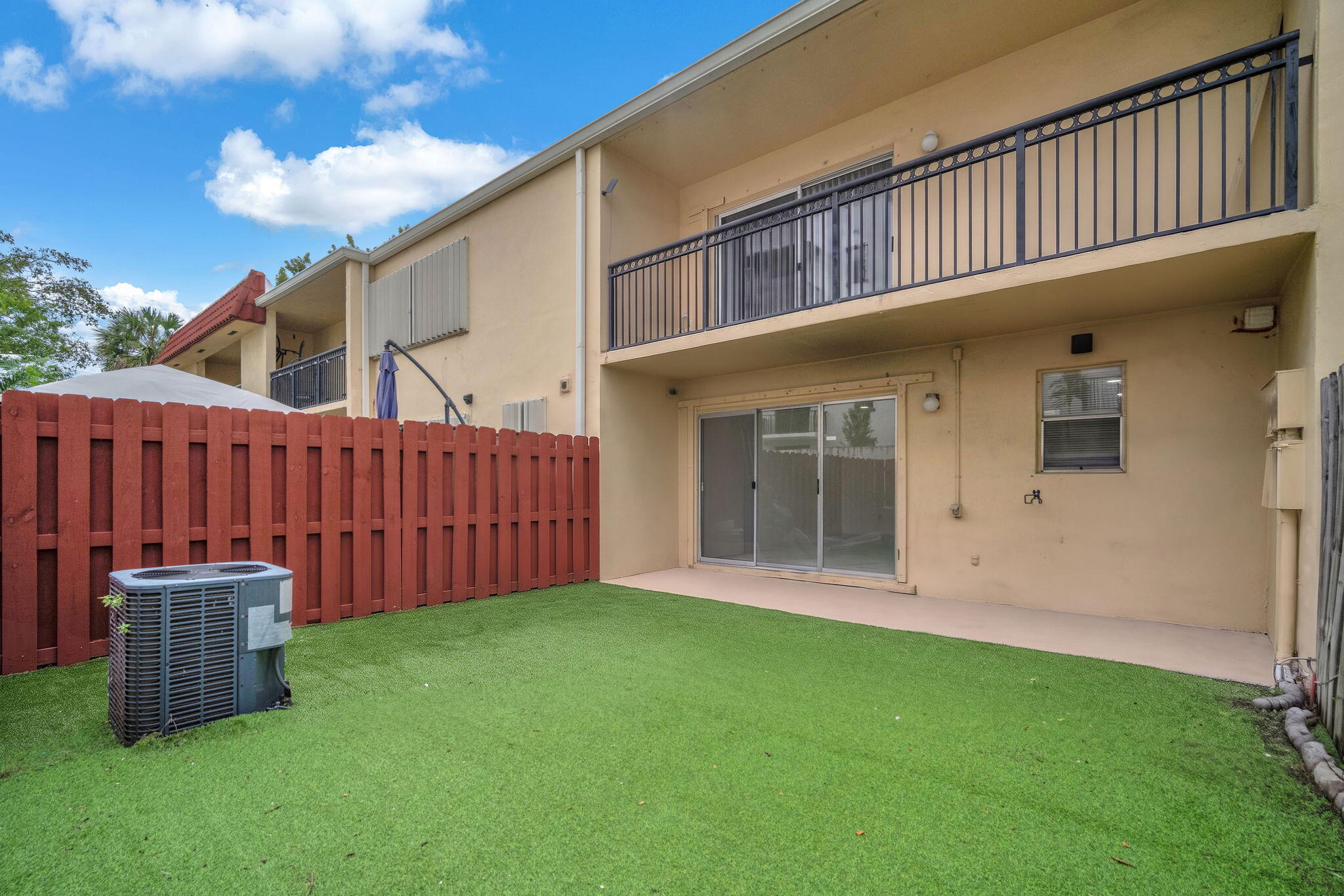 Private backyard with synthetic turf, enclosed by a wood-slat privacy fence, featuring a concrete patio, sliding glass doors, and a second-story balcony with metal railings