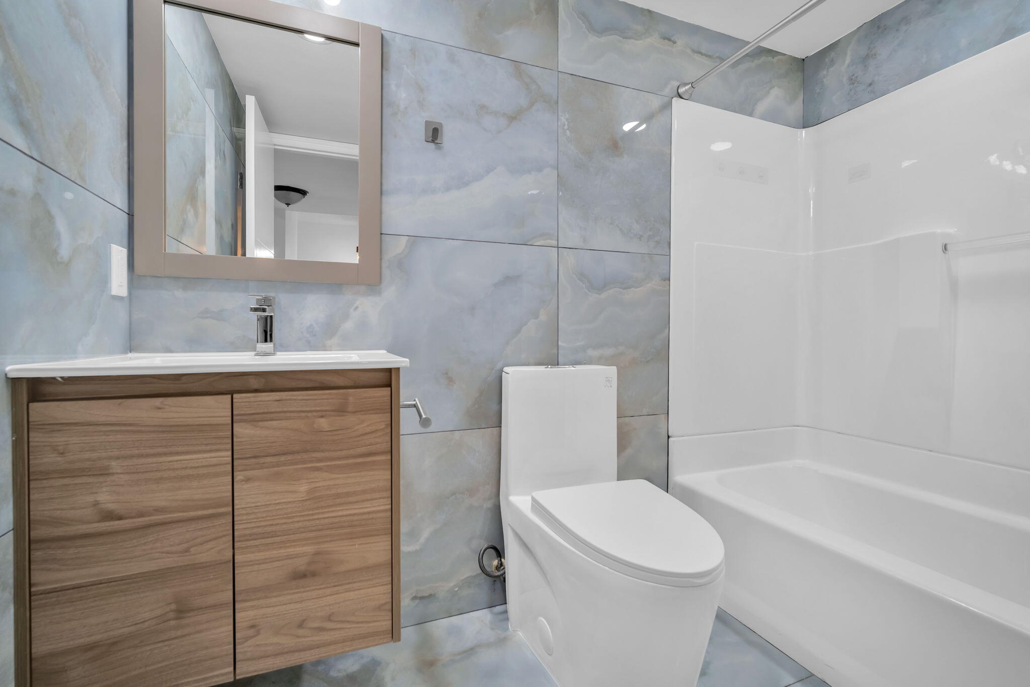 Full-height wall tiling with large-format blue-toned decorative stone-finish panels, wall-mounted wood-finish vanity with a rectangular basin, polished chrome faucet, and white acrylic tub and shower surround