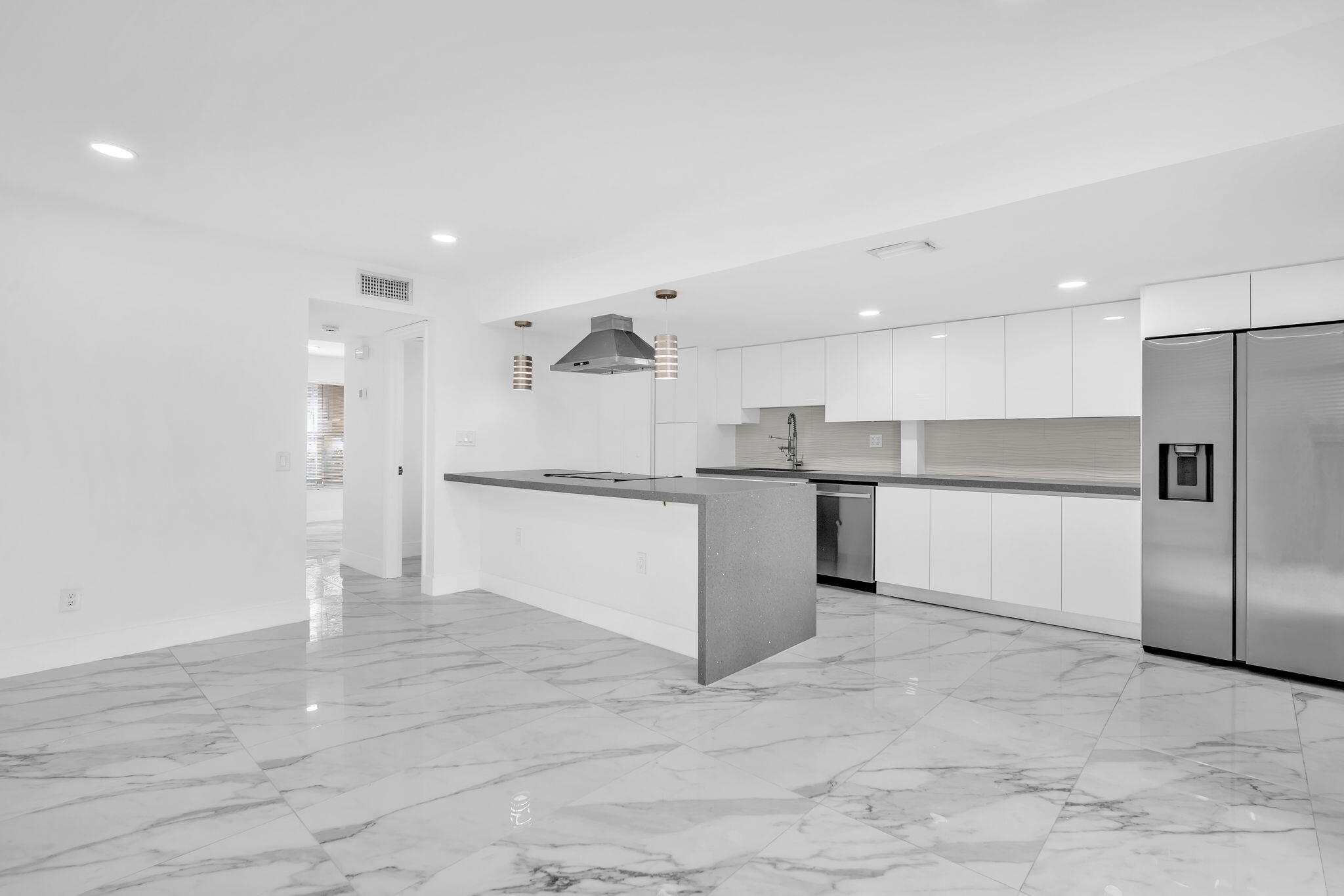 Modern kitchen featuring white flat-panel cabinetry, a waterfall-edge island with an integrated cooktop, polished stone-finish floor tiling, recessed lighting, and stainless steel appliances