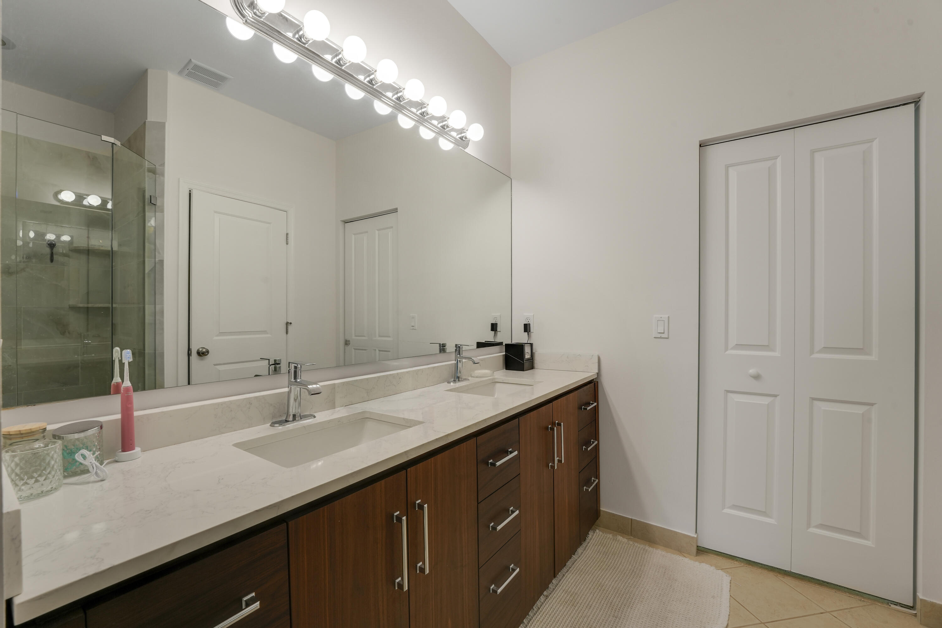 Dual-sink vanity with stone-finish countertop, dark wood-finish cabinetry, full-width mirror, and glass-enclosed shower stall