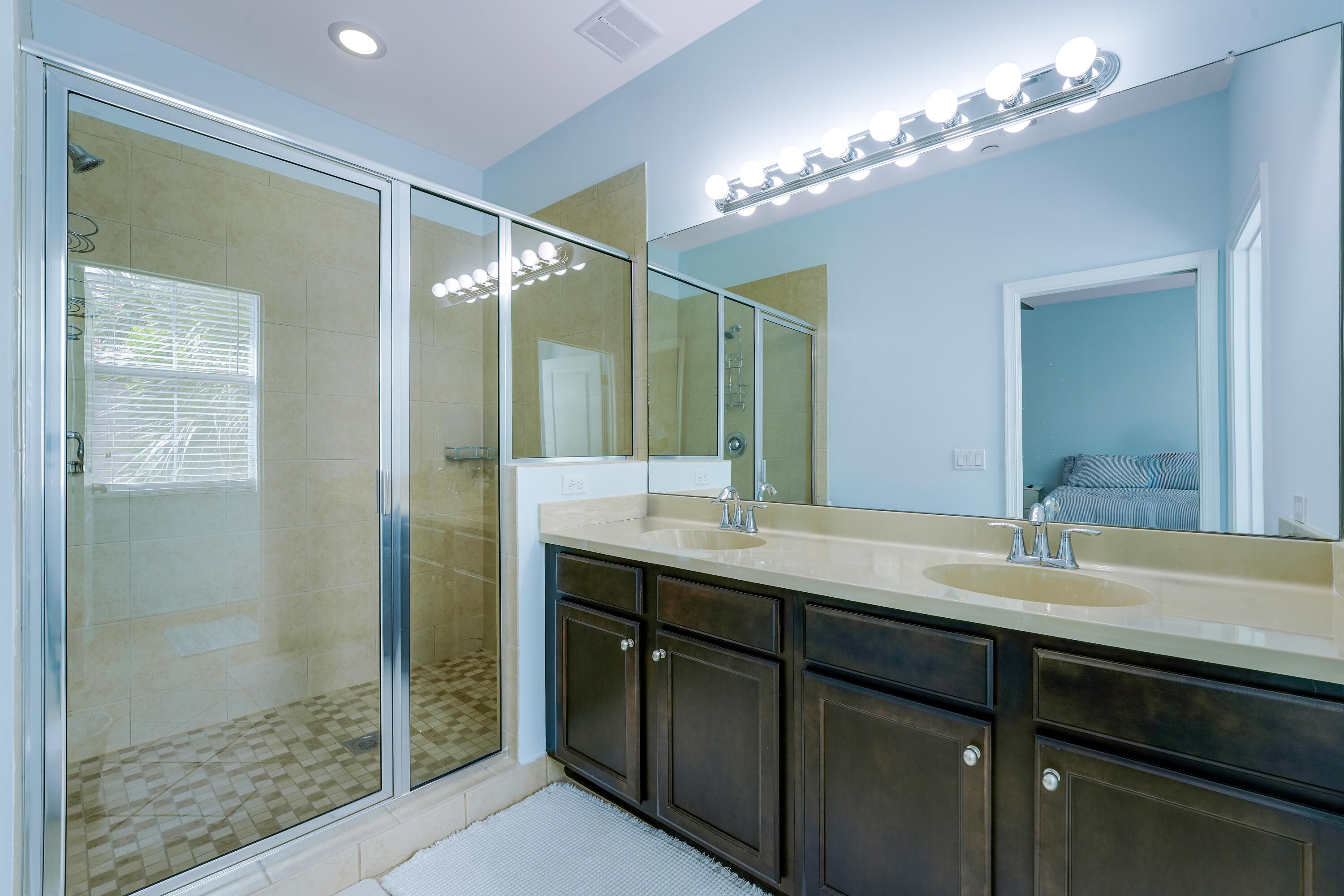 Dual-sink vanity with a solid-surface countertop, floor-to-ceiling mirror, recessed lighting, and a walk-in glass-enclosed shower featuring tile flooring