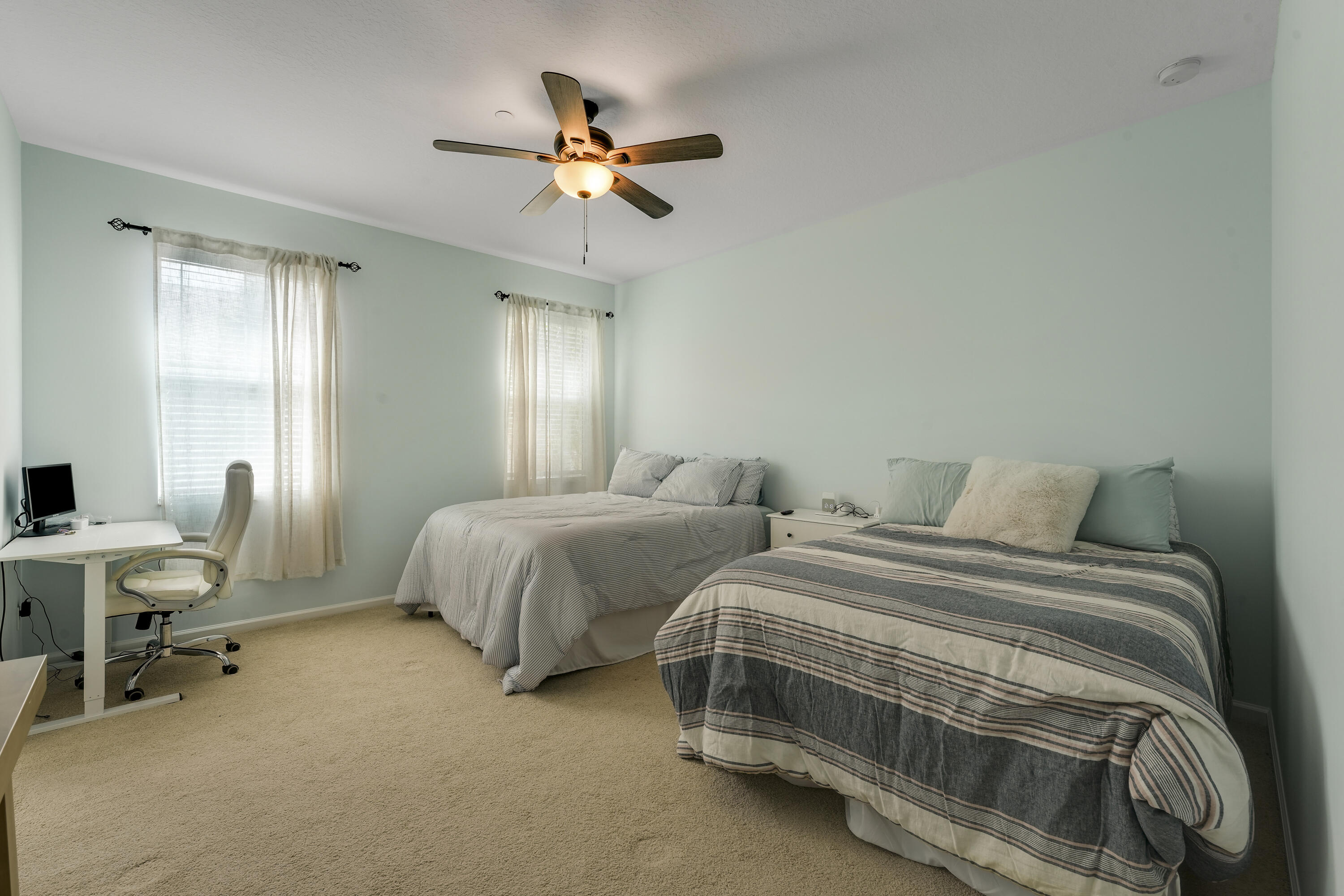 Neutral-toned interior featuring wall-to-wall carpeting, dual windows with installed curtain rods, and a ceiling fan with integrated lighting
