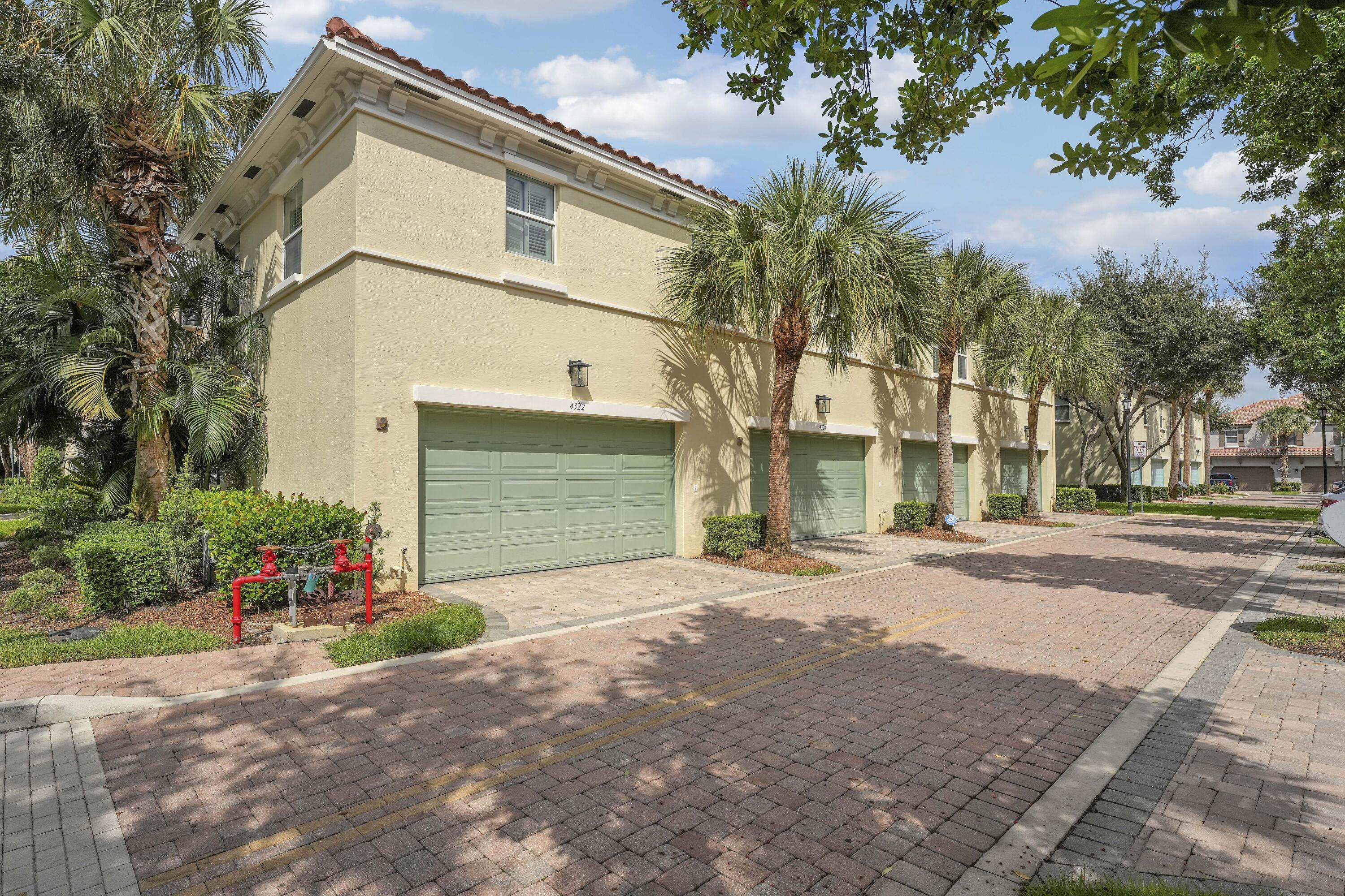 Stucco exterior featuring multiple garage bays, a terracotta-style tile roof, and brick paver driveway, surrounded by mature palm trees