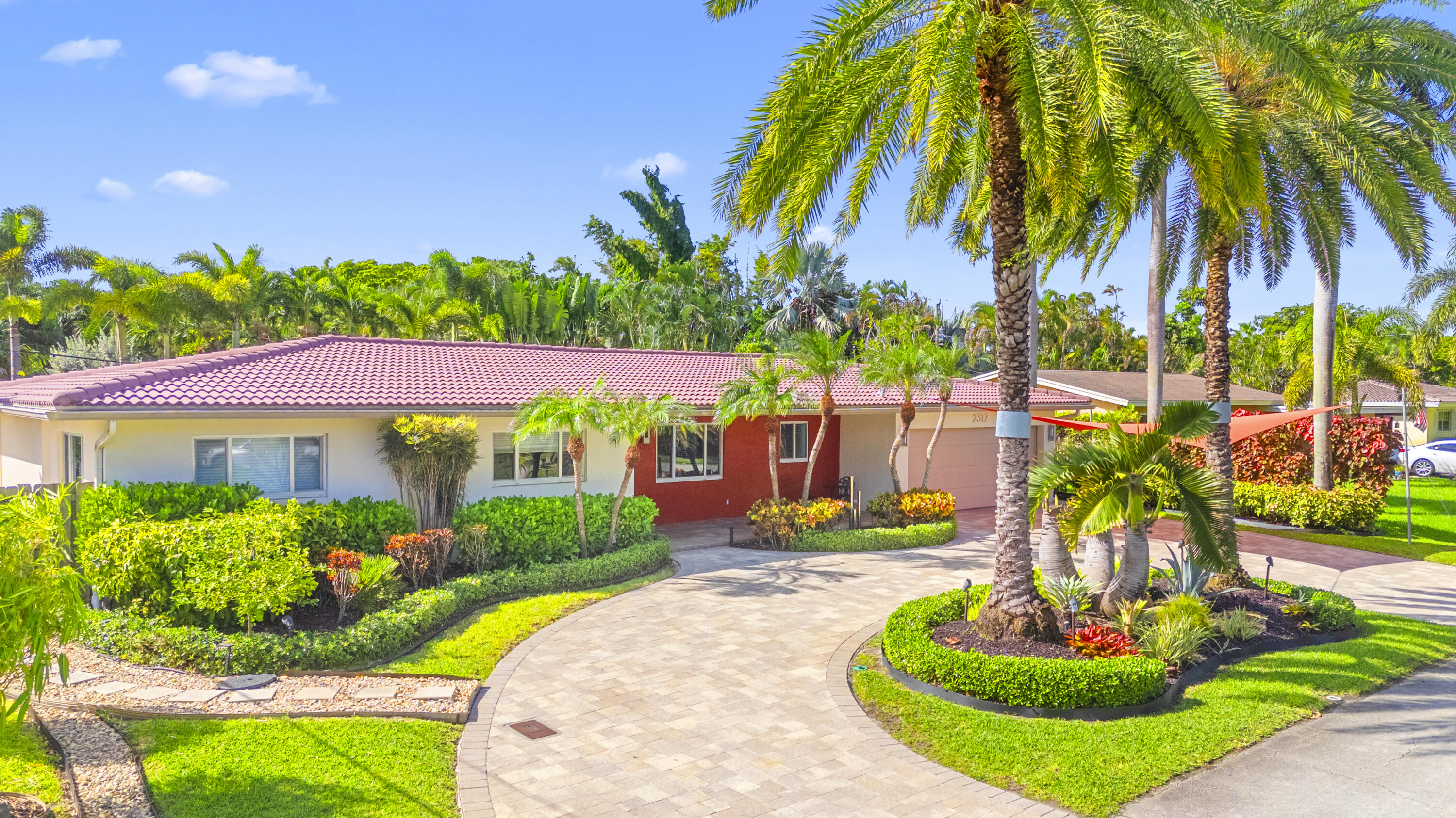 Single-story residence featuring a tile roof, paved circular driveway, manicured landscaping, and mature palm trees