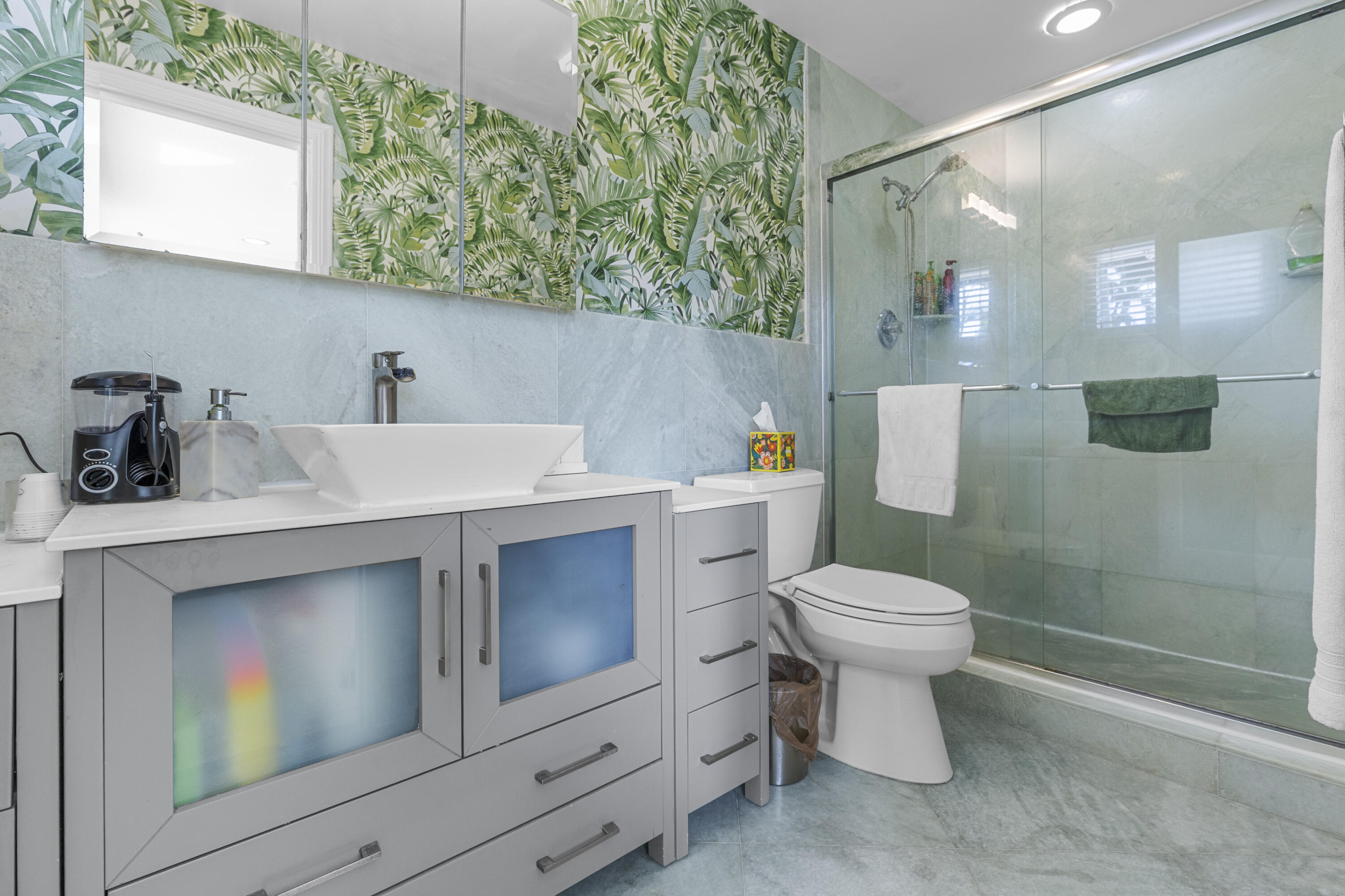 Bathroom featuring a grey vanity with frosted glass cabinet doors, a rectangular vessel sink, stone-finish wall tile, a walk-in shower with a glass enclosure, and patterned wallpaper