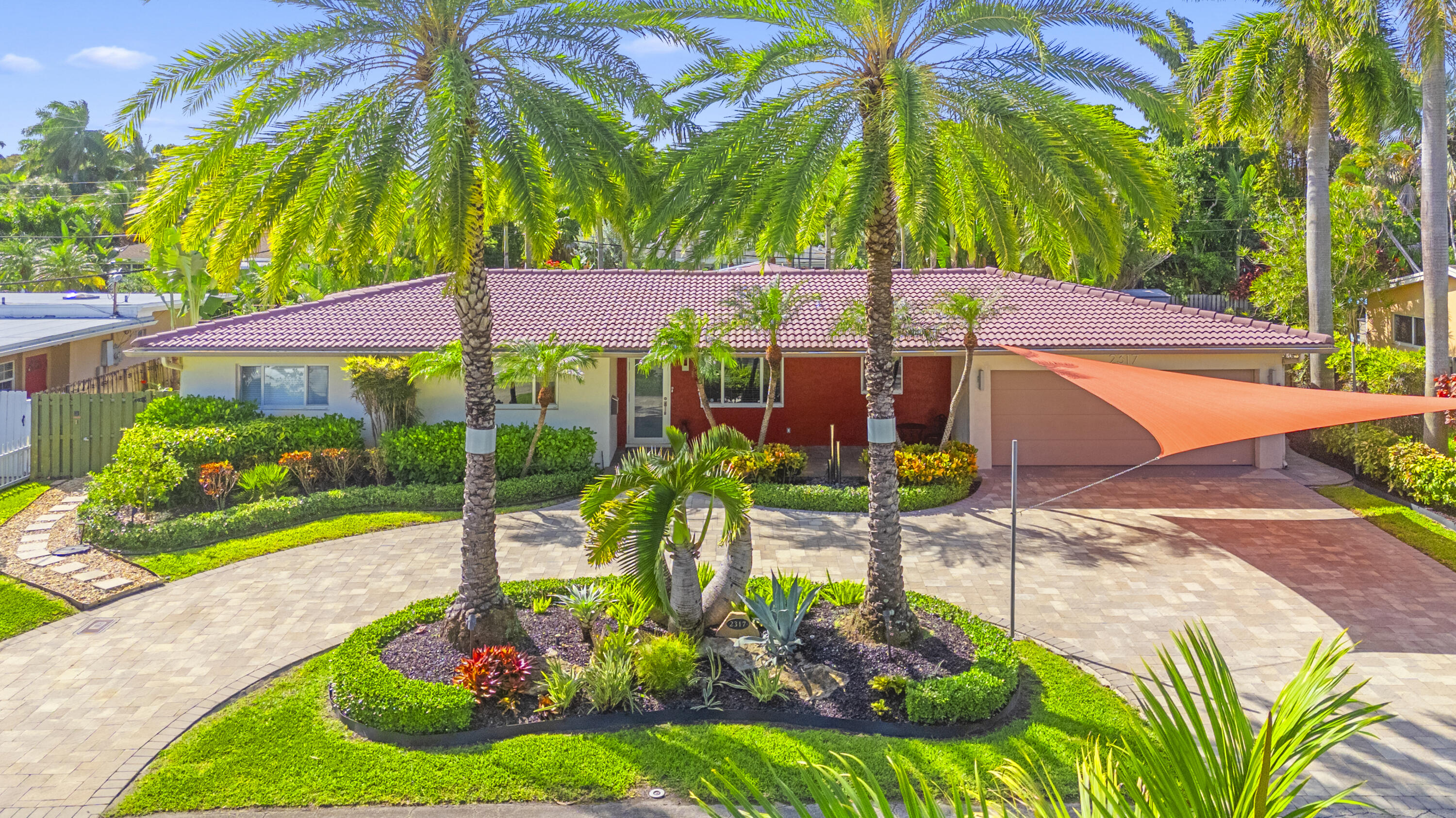 Single-story residence featuring a tile roof, paved circular driveway, mature palm trees, professional landscaping, and a two-car garage