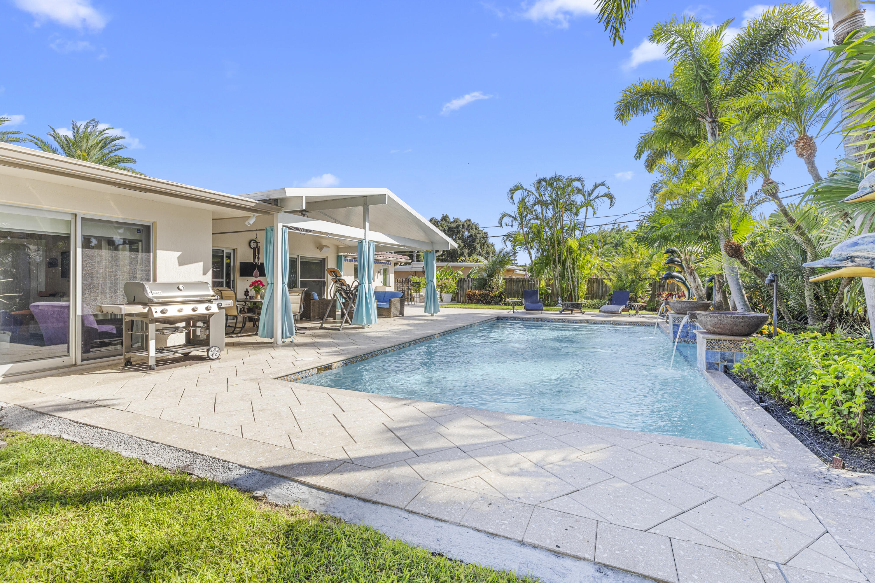 Outdoor living space featuring a covered patio, swimming pool with water features, patterned paver decking, and mature palm trees