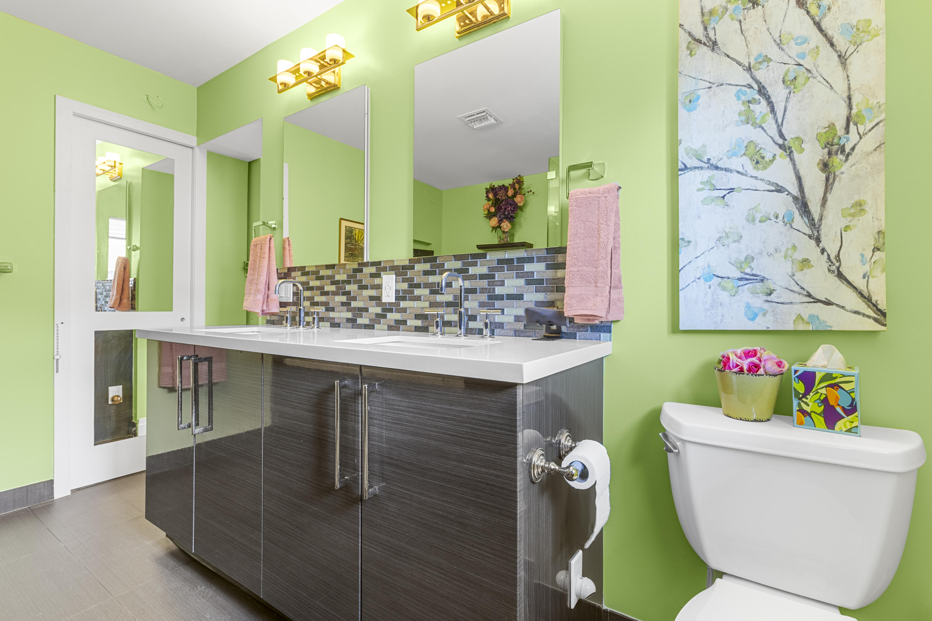 Dual-sink vanity featuring a solid surface countertop, high-gloss cabinetry, linear tile backsplash, and wall-mounted lighting