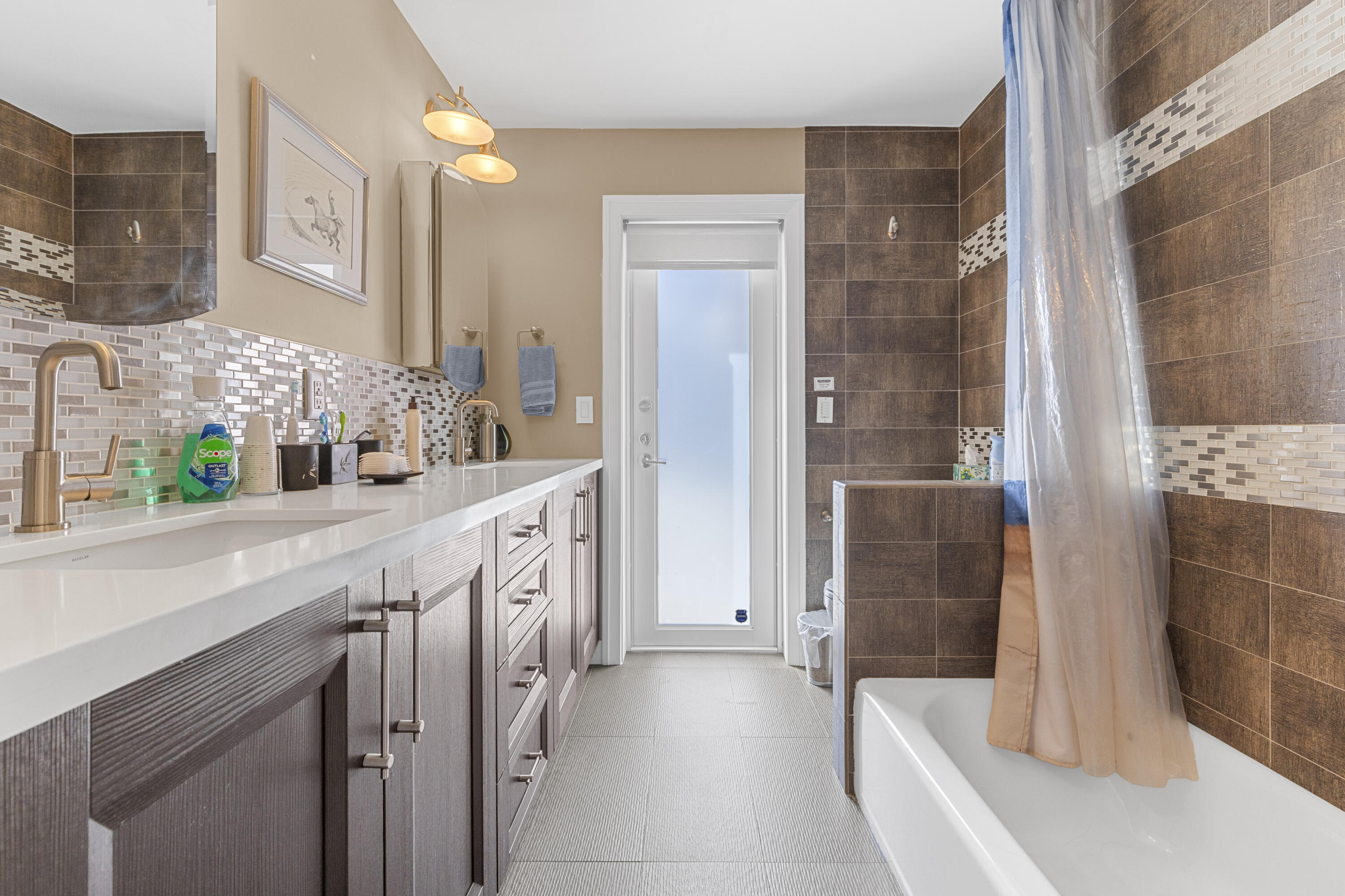 Bathroom featuring custom wood-finish cabinetry, white solid-surface countertop, metallic finish fixtures, glass tile backsplash, and a tiled shower surround