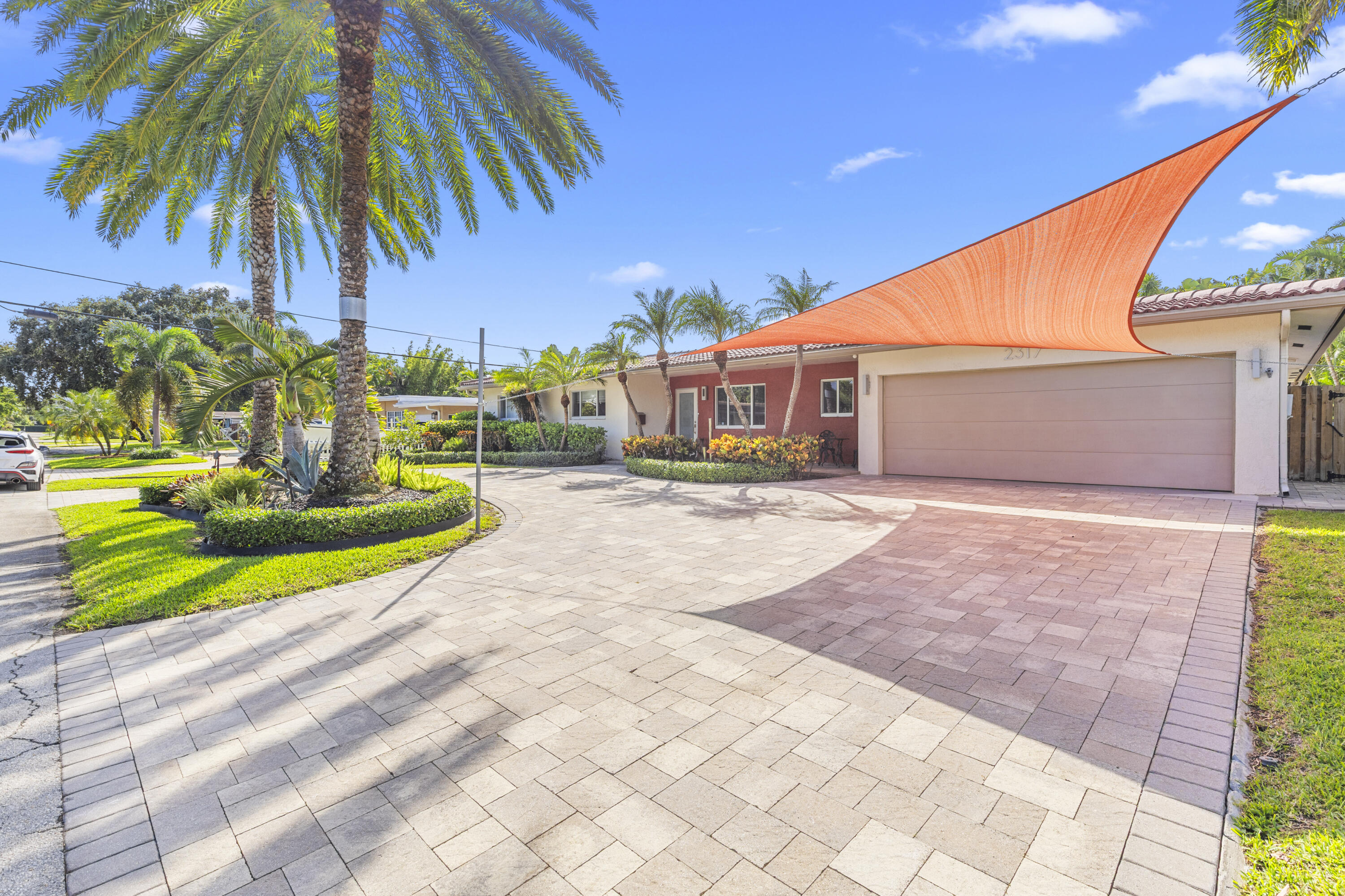 Exterior facade featuring a two-car garage, paver driveway, tiled roof, mature palm trees, and manicured landscaping