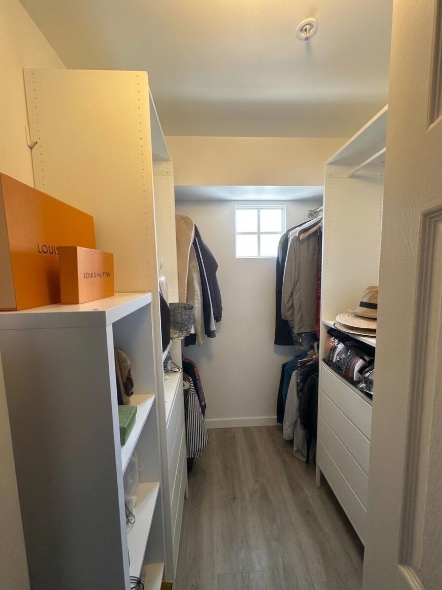 Walk-in closet featuring white built-in shelving, integrated storage drawers, hanging rods, wood-finish flooring, and a single-pane window