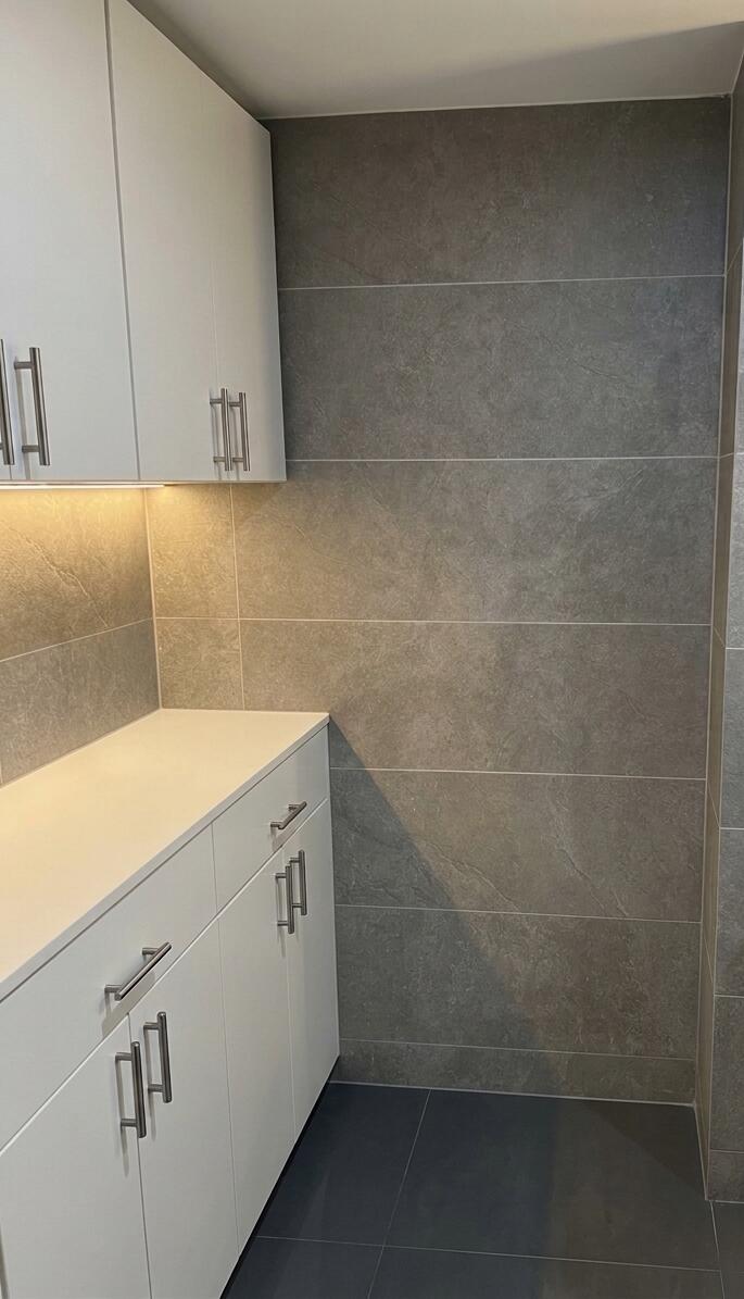 Built-in white cabinetry featuring metallic hardware, solid-surface countertop, integrated under-cabinet lighting, and large-format grey tile wall cladding