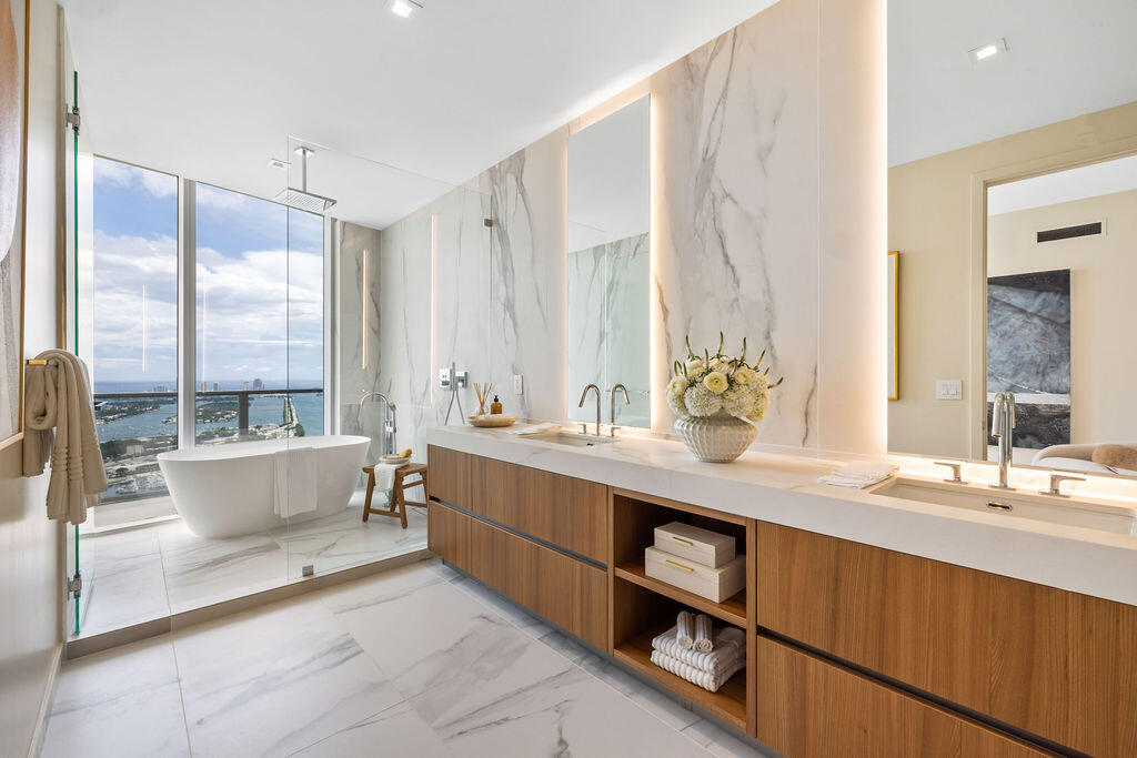 Bathroom featuring floor-to-ceiling stone wall cladding, a floating wood-finish dual vanity with integrated countertop basins, a deep soaking tub, and expansive floor-to-ceiling windows