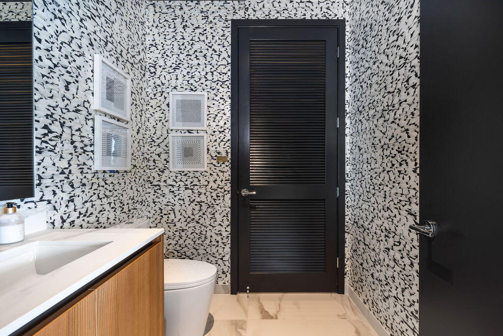 High-contrast patterned wall tile, vanity with a rectangular sink, light-toned countertop, black louvered door, and large-format neutral tile flooring