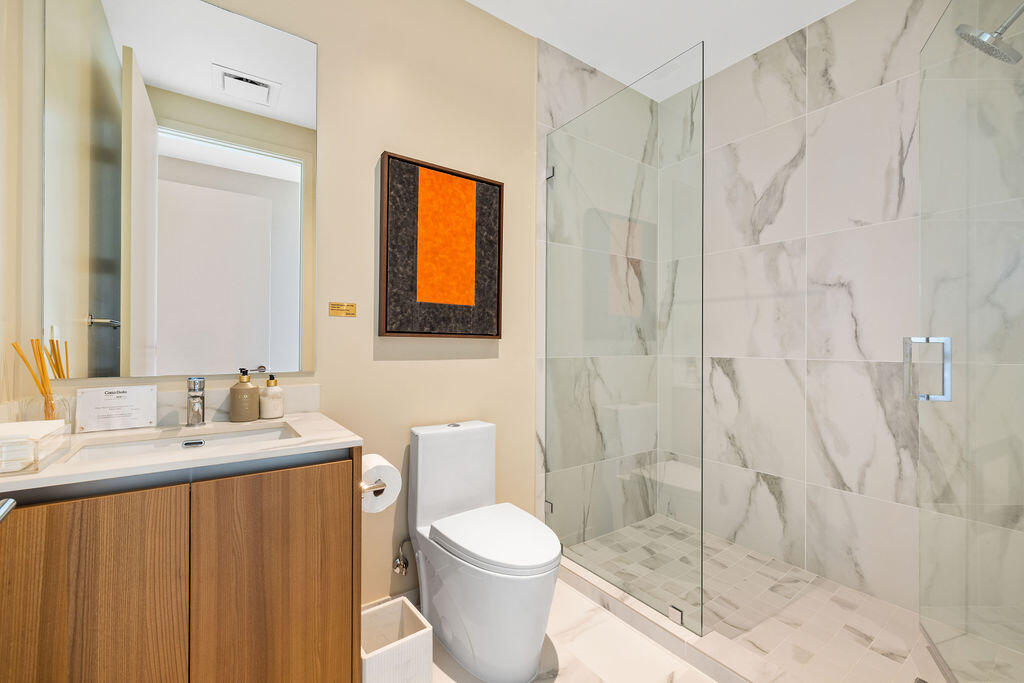Bathroom featuring a wood-finish vanity with a stone countertop, wall-to-wall mirror, glass-enclosed walk-in shower with floor-to-ceiling stone-style tile, and a dual-flush toilet