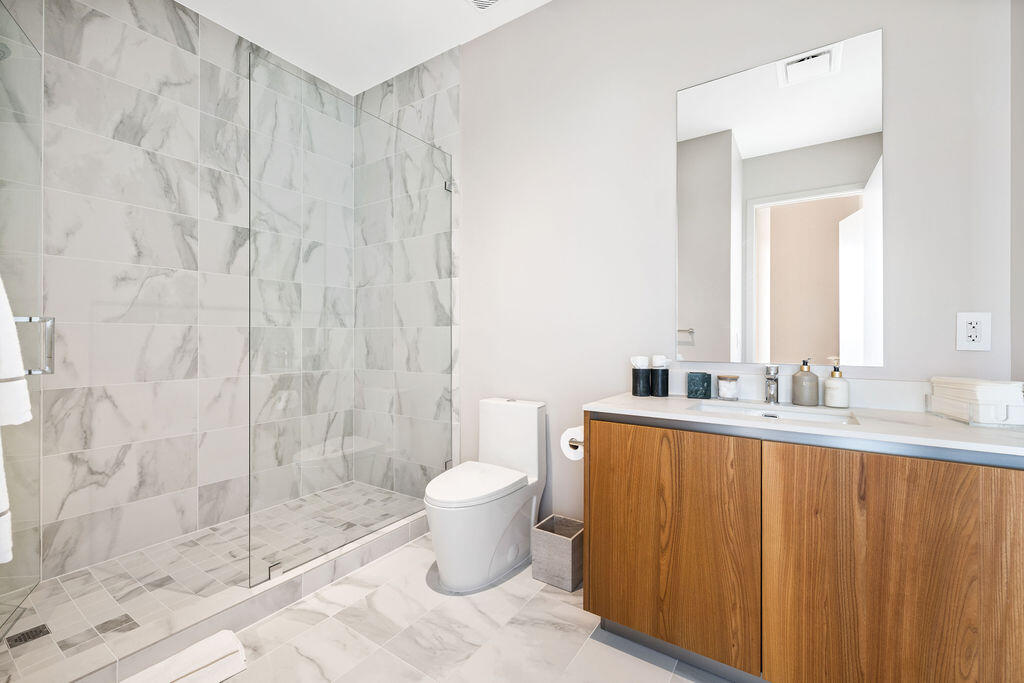 Bathroom featuring a walk-in shower with frameless glass enclosure, floor-to-ceiling stone-pattern tile, wood-finish cabinetry, white countertop, and frameless wall-mounted mirror