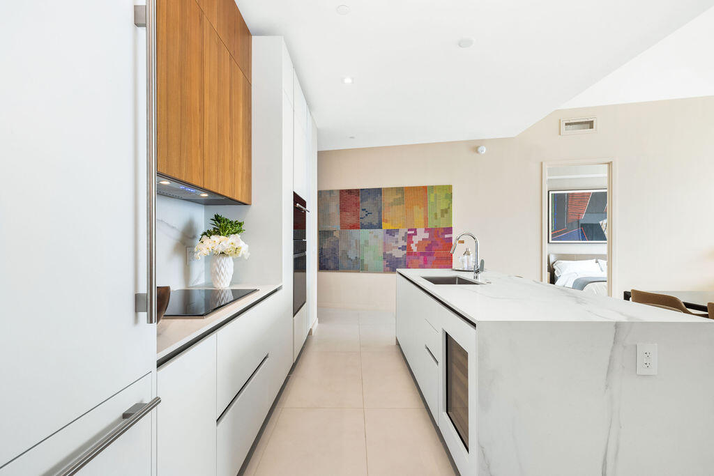 Kitchen featuring white flat-panel cabinetry, wood-finish upper cabinets, stone-surfaced waterfall island, built-in wall oven, and integrated wine cooler