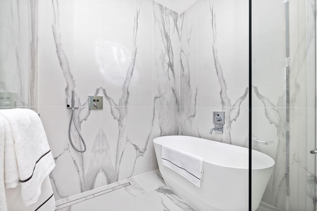 Freestanding soaking tub featuring a wall-mounted faucet, floor-to-ceiling large-format stone-finish tile surround, handheld shower fixture, and glass shower enclosure