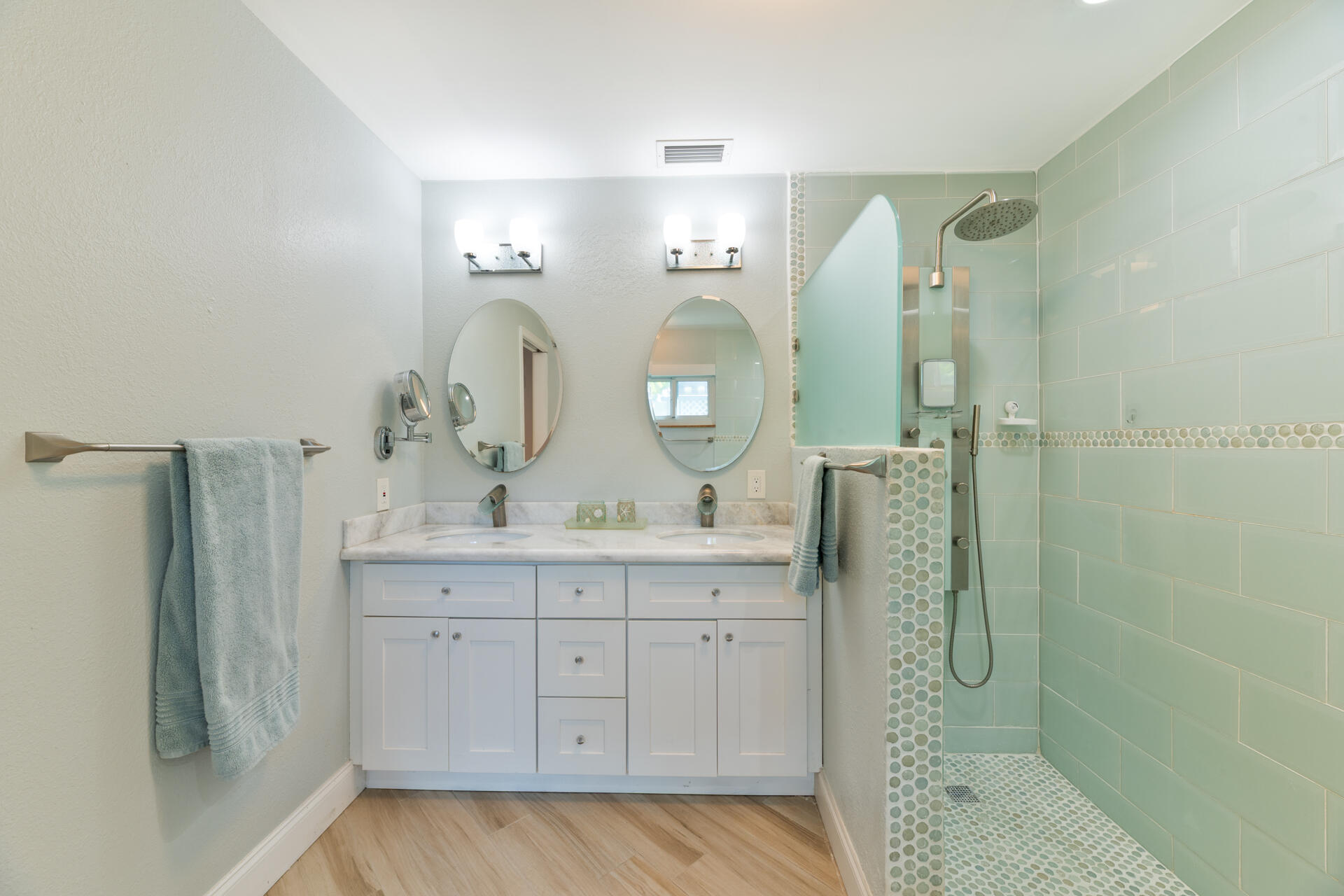 Dual-sink vanity with white cabinetry and stone countertop, dual oval wall mirrors, walk-in shower featuring glass tile surround, and wood-finish flooring