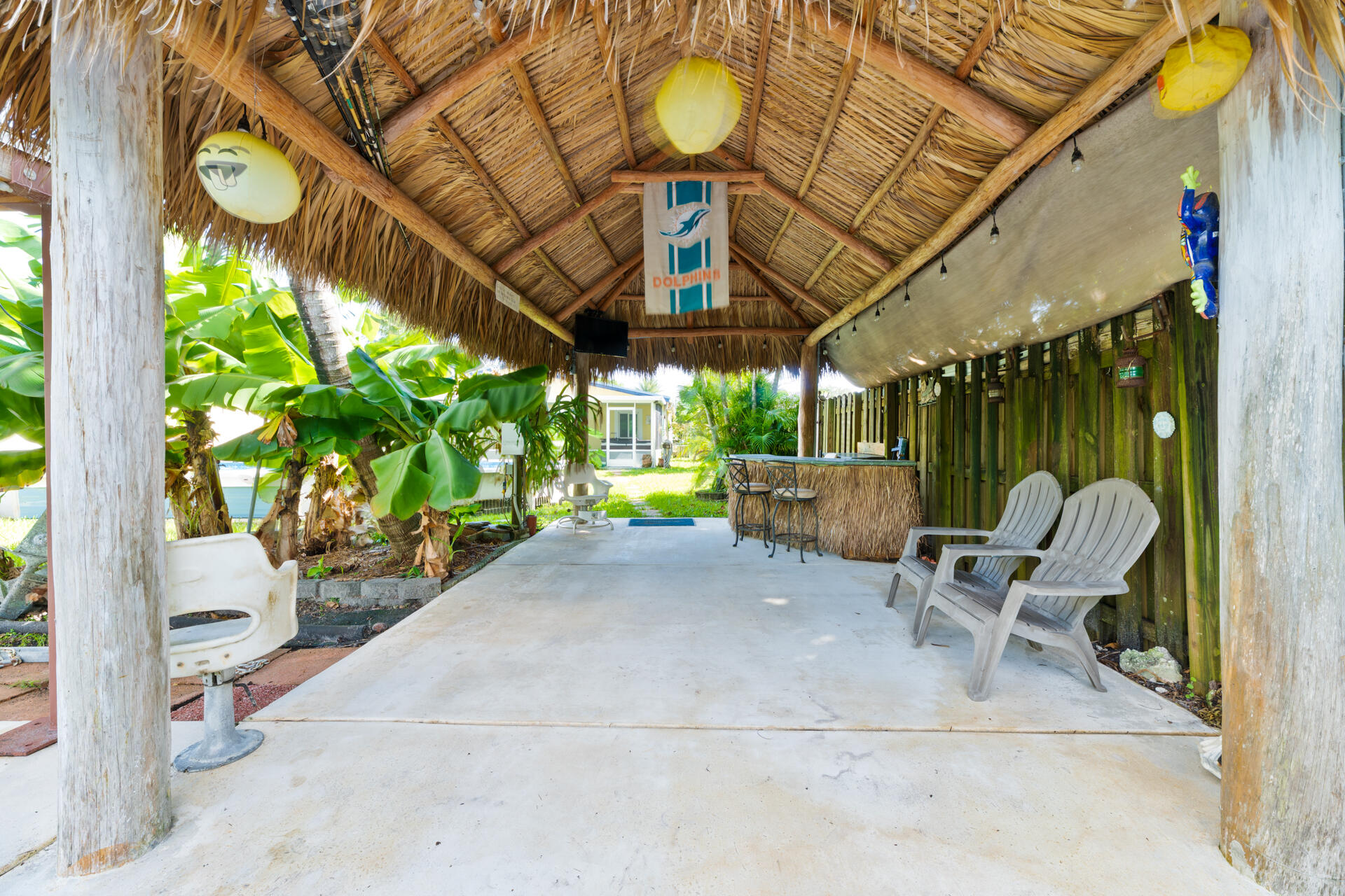 Covered outdoor lounge featuring a thatched palm roof, exposed timber support columns, concrete flooring, and a stand alone bar