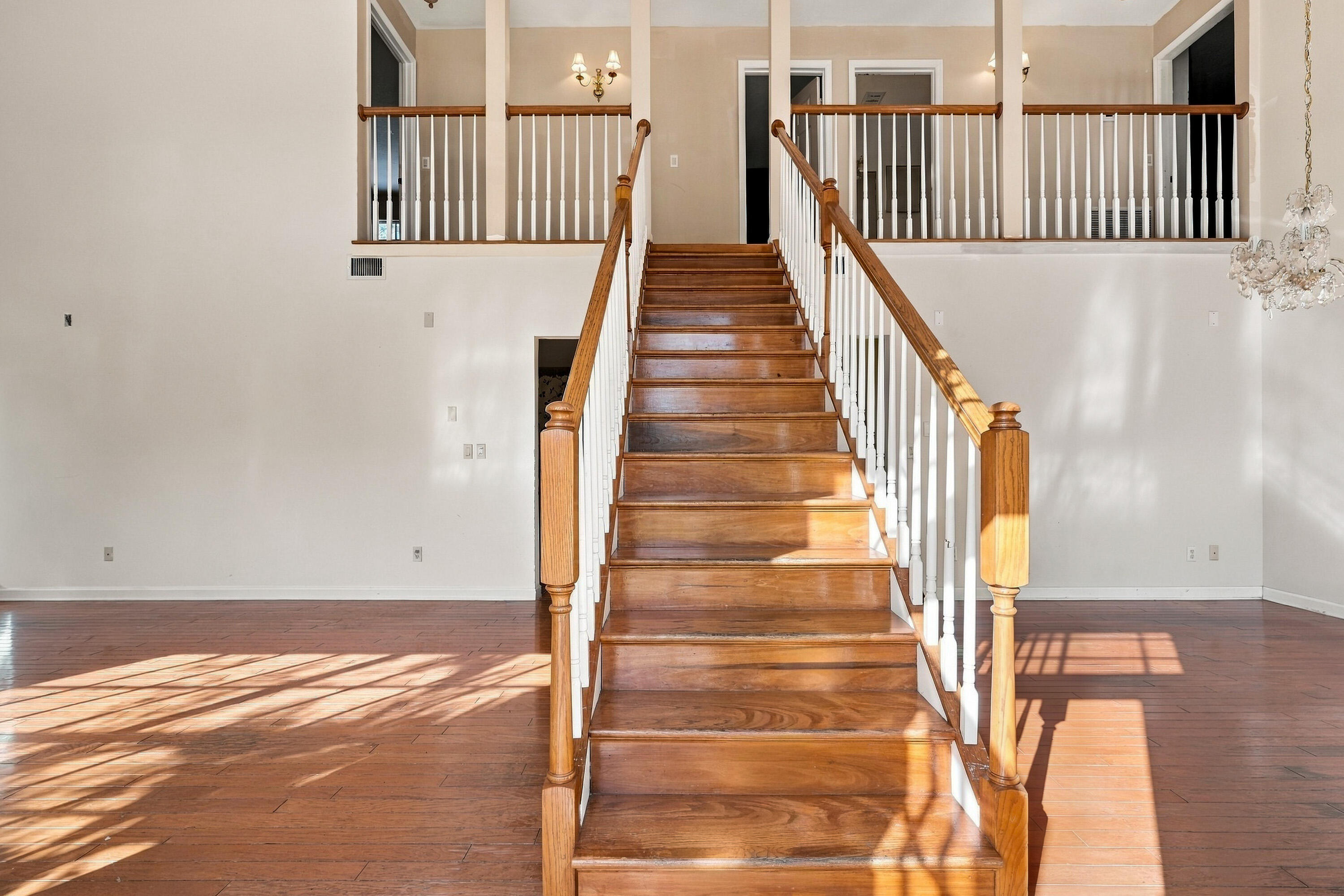 Open foyer featuring a wood-finish staircase with a custom wooden handrail, white vertical balusters, wood-finish flooring, and a decorative glass chandelier Open foyer featuring a wood-finish staircase with a custom wooden handrail, white vertical balusters, wood-finish flooring, and a decorative glass chandelier