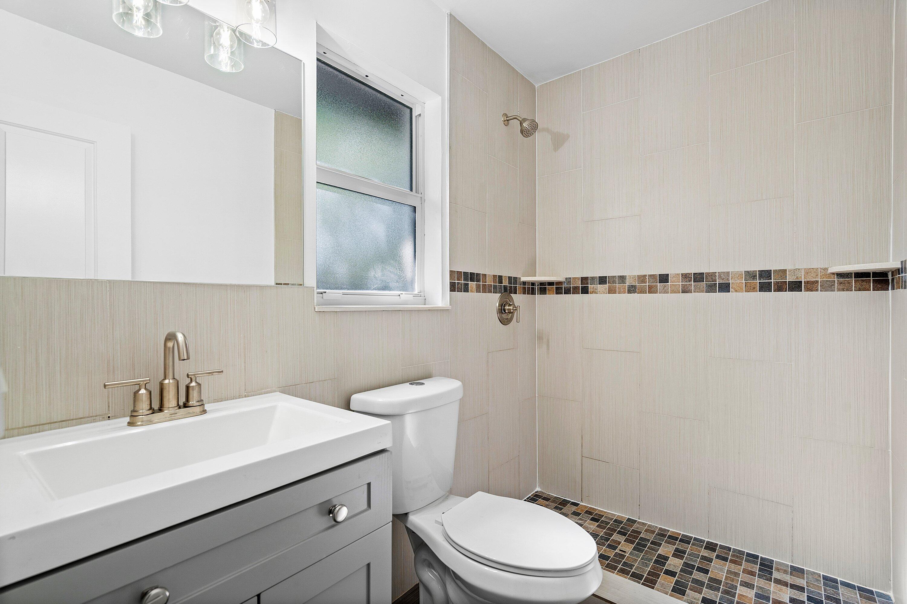 Bathroom featuring a gray vanity with an integrated sink, brushed-metal hardware, a wall-mounted mirror, and a walk-in shower with neutral tile and decorative mosaic accents