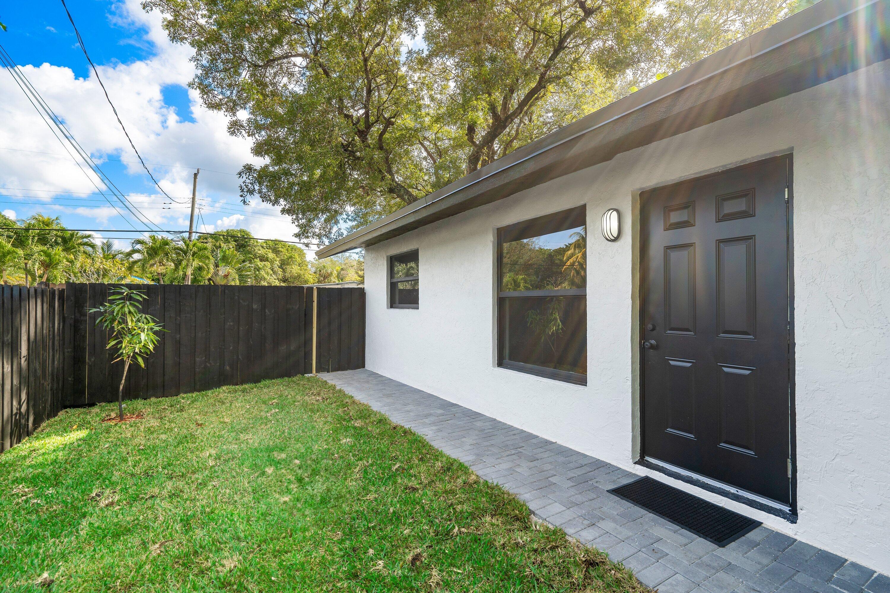 Exterior featuring a white stucco facade, a dark-finished panel door, a grey paver walkway, and a fenced yard with mature trees