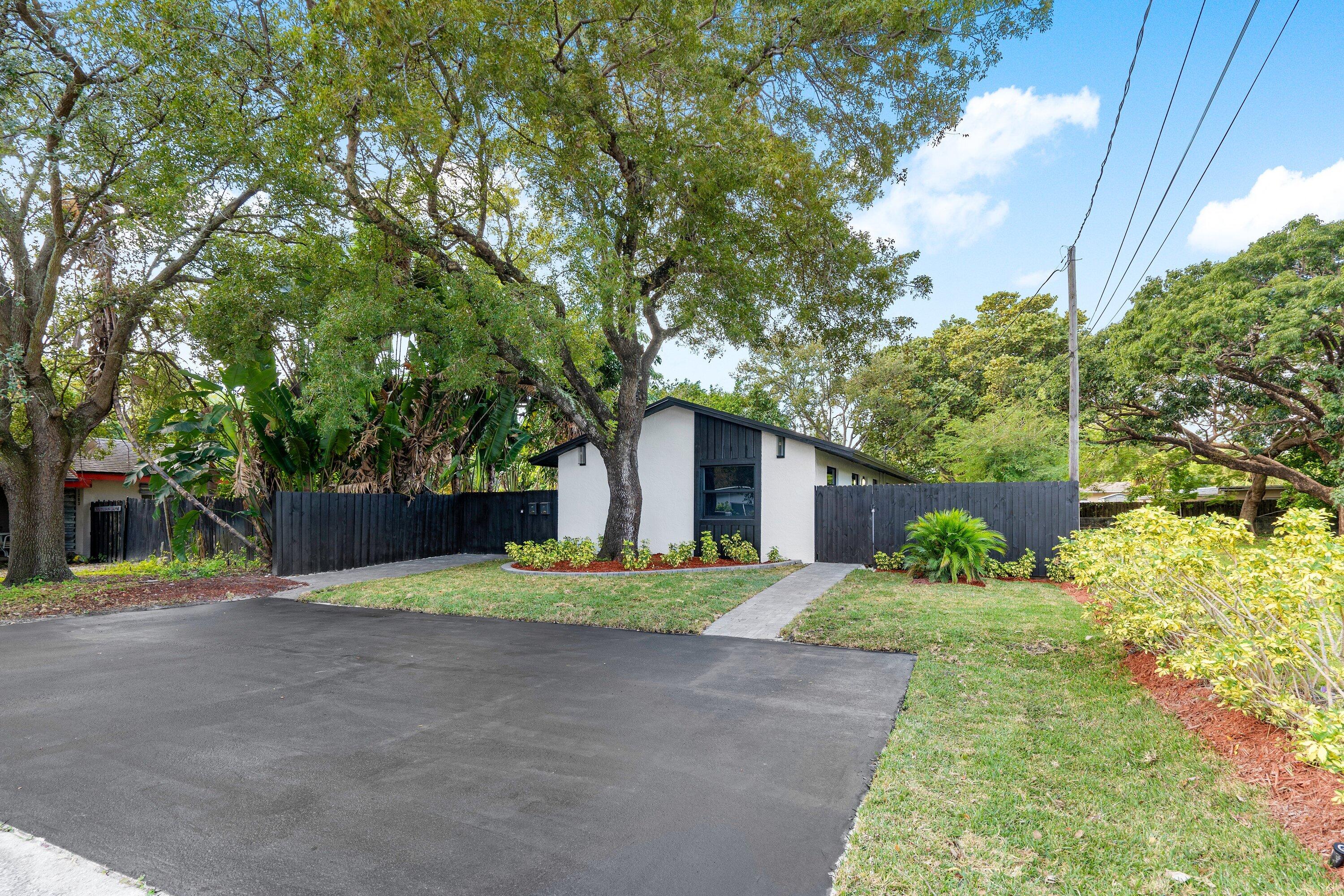 Exterior featuring a white facade with black architectural accents, asphalt driveway, mature trees, and enclosed perimeter fencing