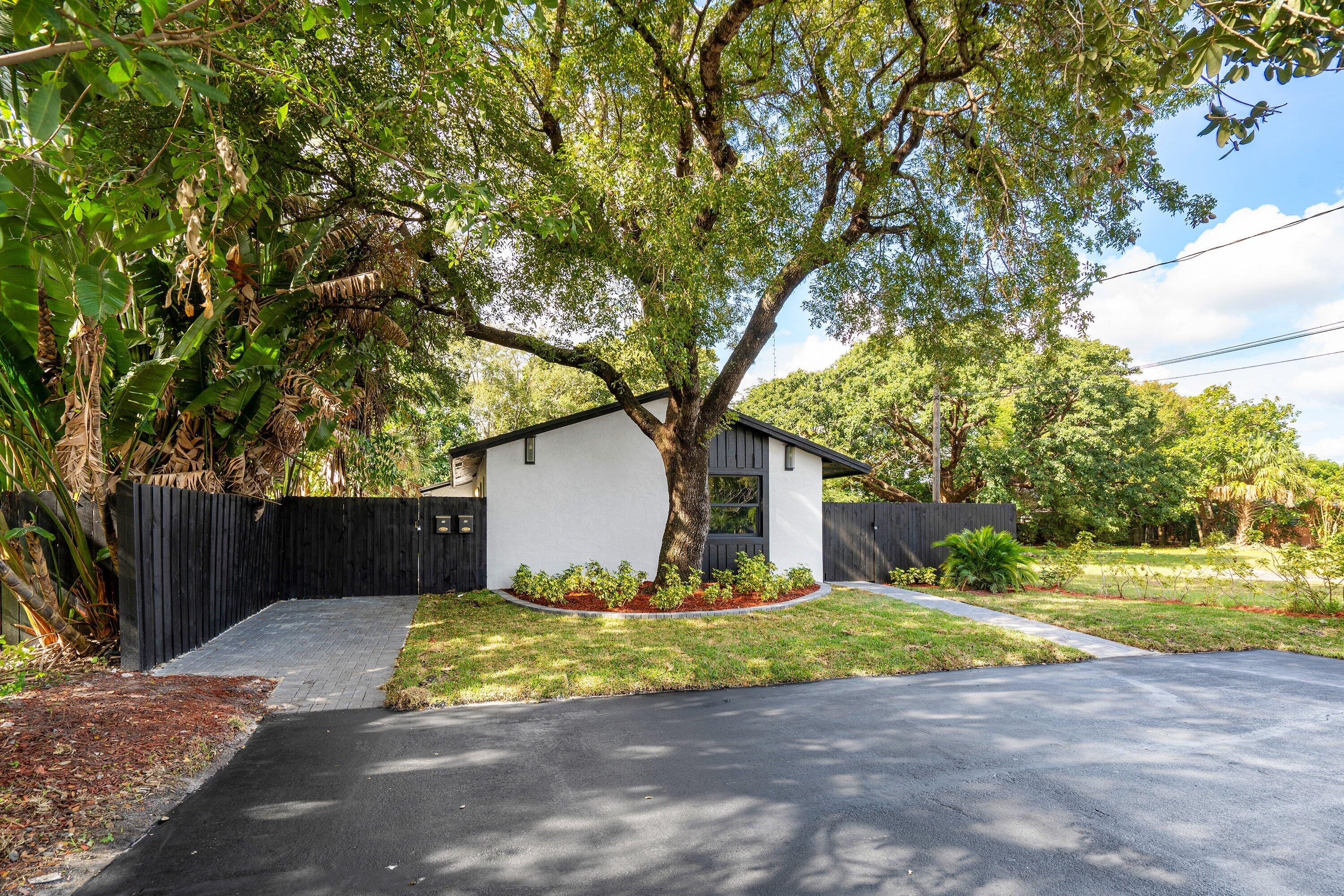 Exterior facade featuring white stucco walls, vertical dark-toned siding, mature tree canopy, stone-paver driveway, and solid dark-toned privacy fencing