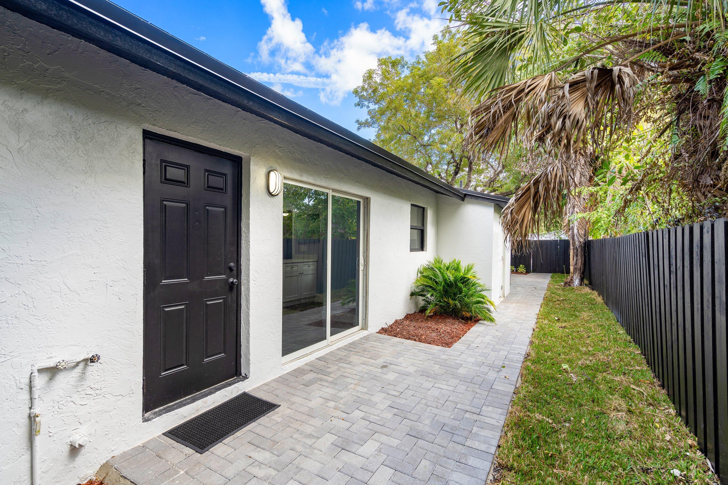 Exterior space featuring white stucco siding, dark-toned panel door, sliding glass door, grey paver patio, and dark-toned perimeter fencing
