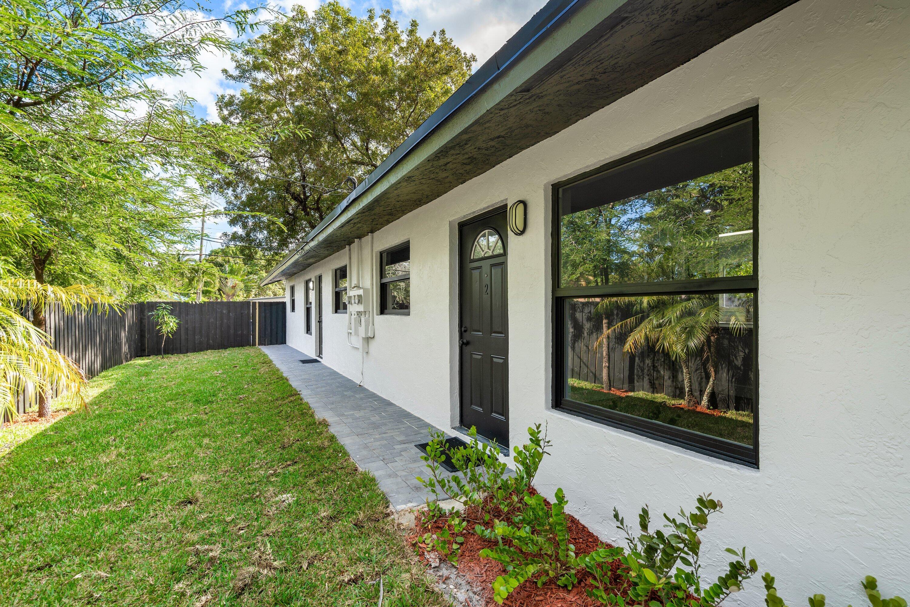 Exterior featuring white stucco siding, dark-toned window frames, paver walkway, and fenced yard space with mature trees Exterior featuring white stucco siding, dark-toned window frames, paver walkway, and fenced yard space with mature trees