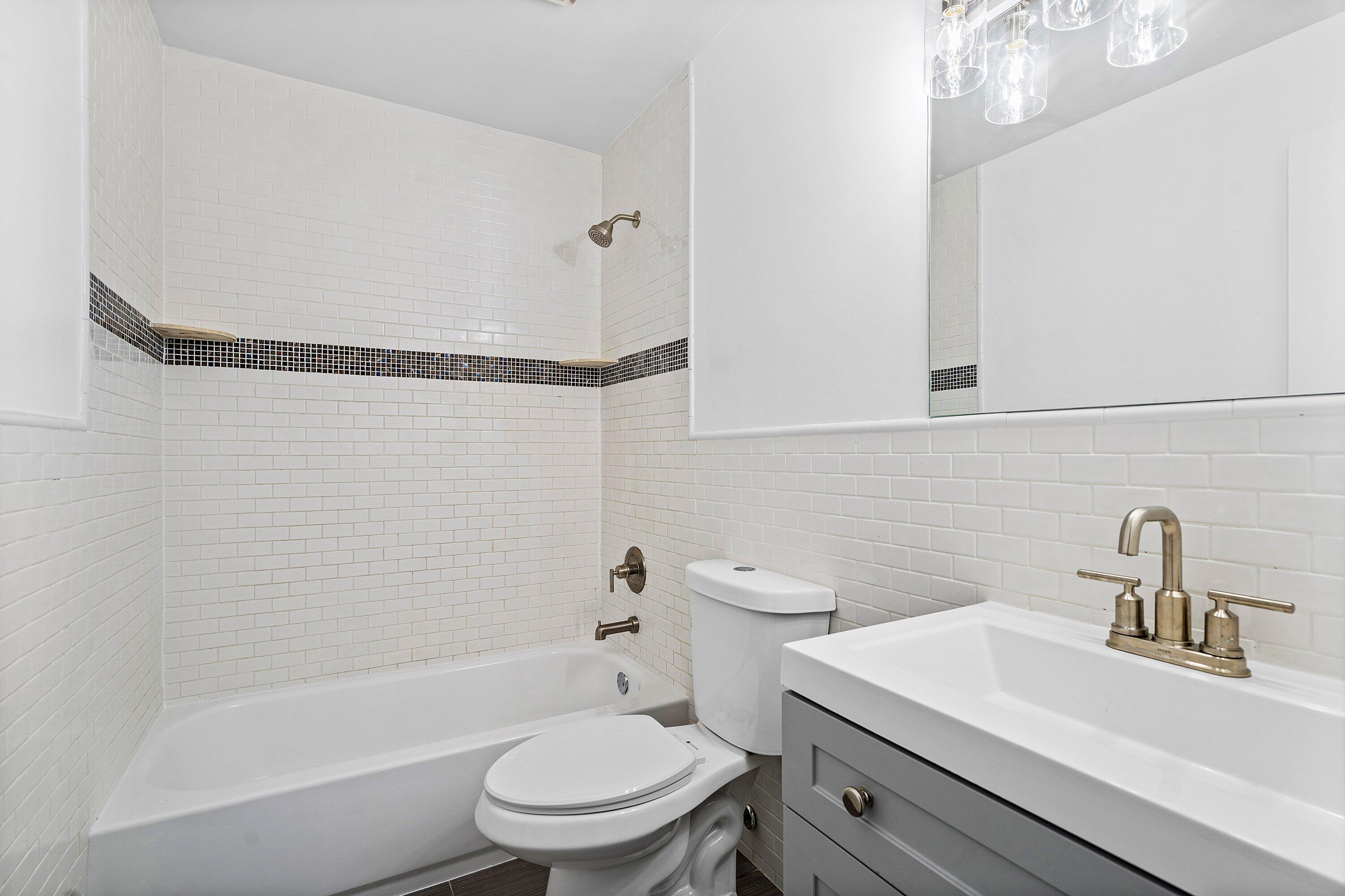 Bathroom featuring white subway tile walls with a mosaic tile inlay, a soaking tub with a wall-mounted showerhead, a grey vanity with a rectangular integrated sink, and metallic finish plumbing fixtures