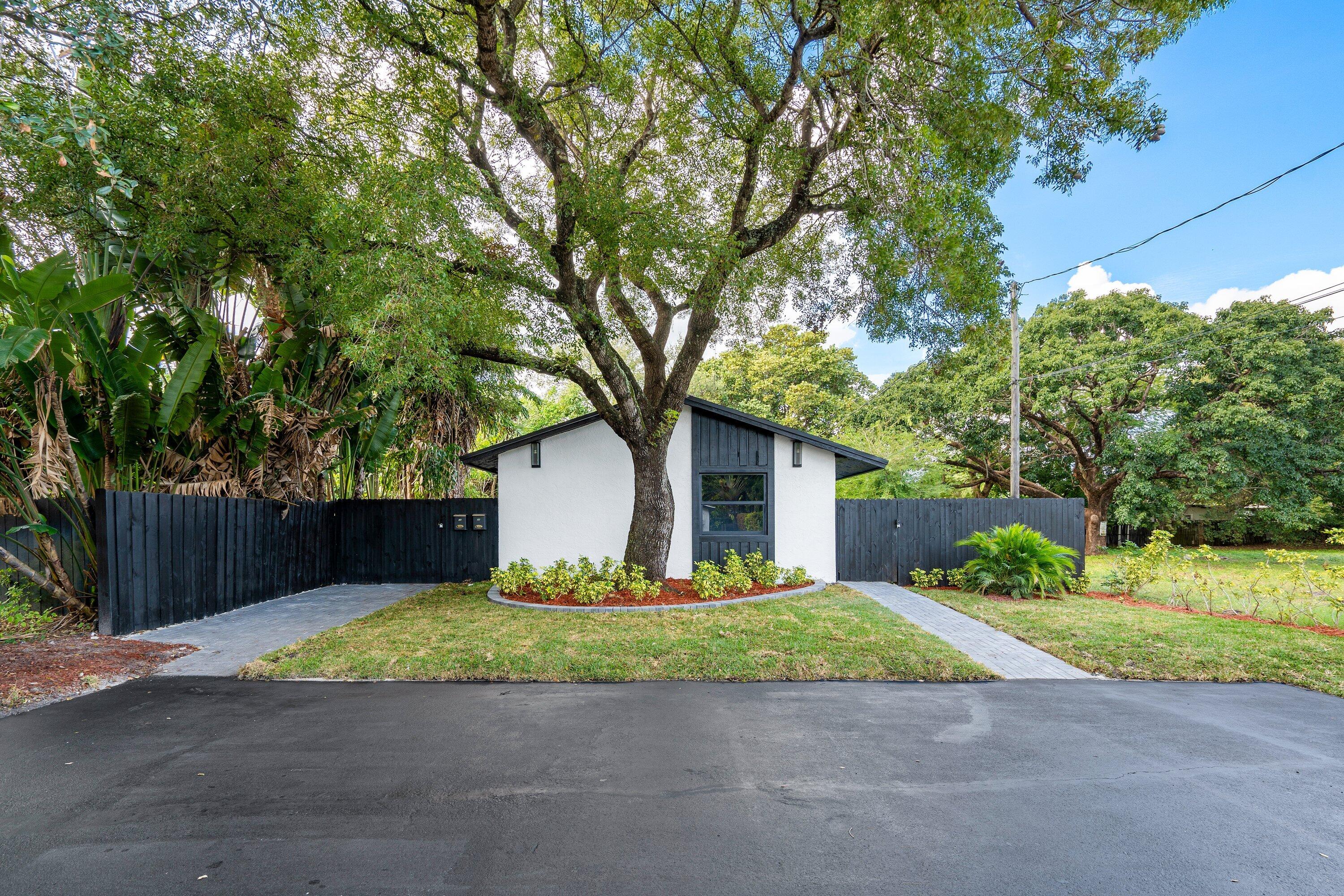 Exterior facade featuring white siding with dark-toned trim, asphalt-paved driveway, decorative landscaping with a central mature tree, and dark wood-style privacy fencing