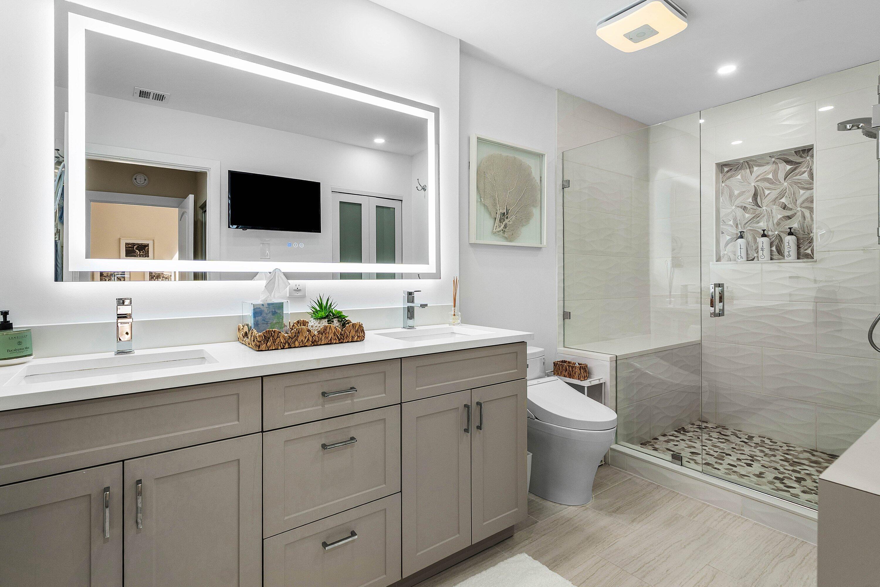 Dual-vanity cabinet with quartz countertop, wall-to-wall LED-backlit mirror, glass-enclosed shower with decorative tile inlay, and stone-tiled shower flooring