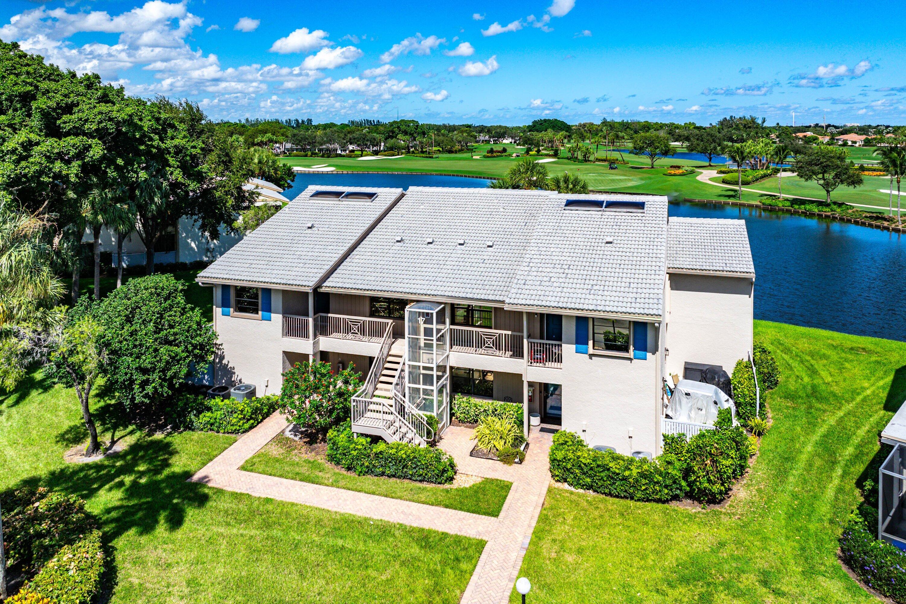 Two-story building featuring a tile roof with integrated solar panels, an exterior glass-enclosed elevator, balconies with railings, and manicured landscaping situated on a water-front lot