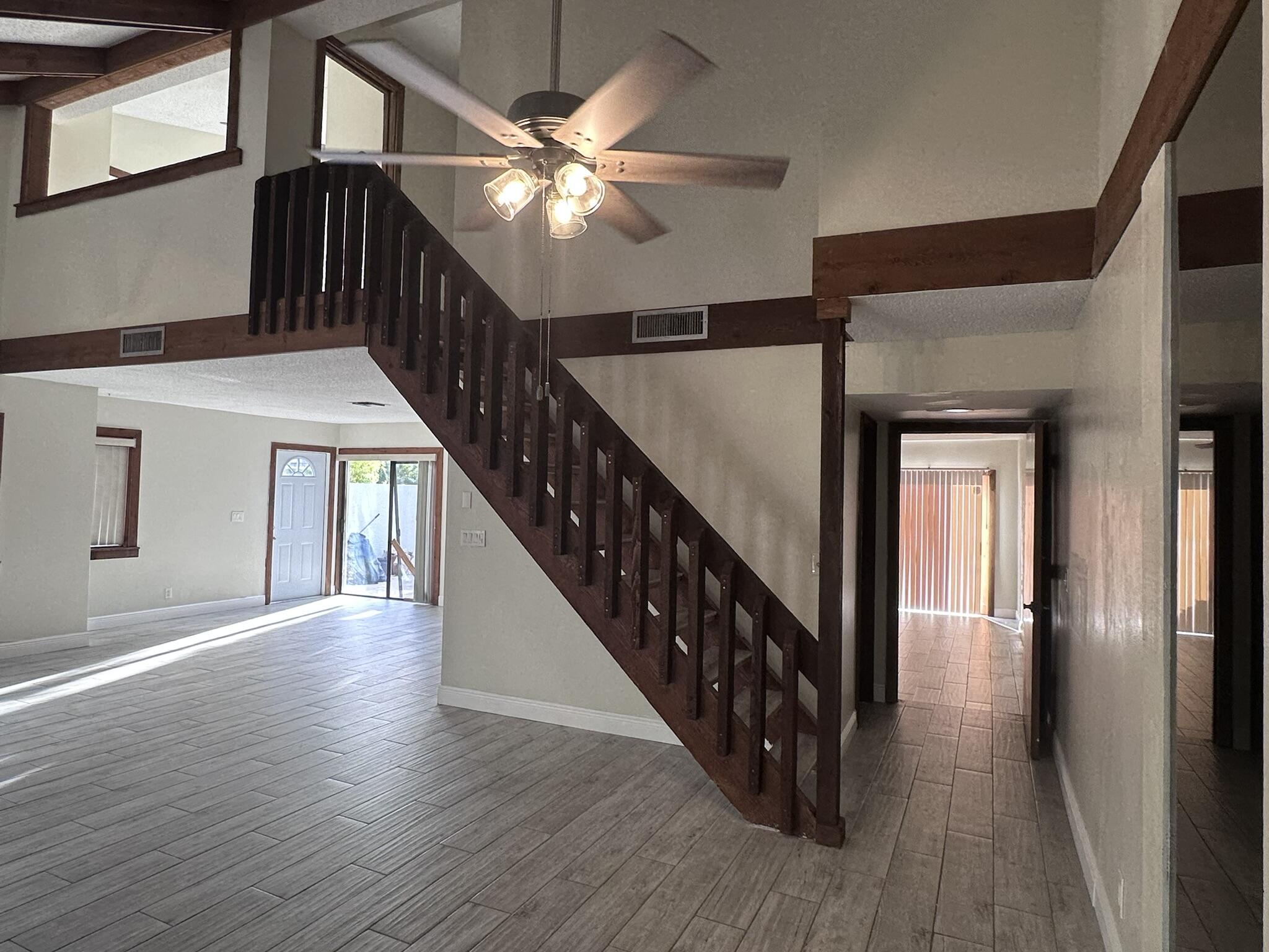Open-concept interior featuring vaulted ceilings with exposed wood beams, wood-finish tile flooring, a prominent staircase with dark wood railings, and a multi-light ceiling fan