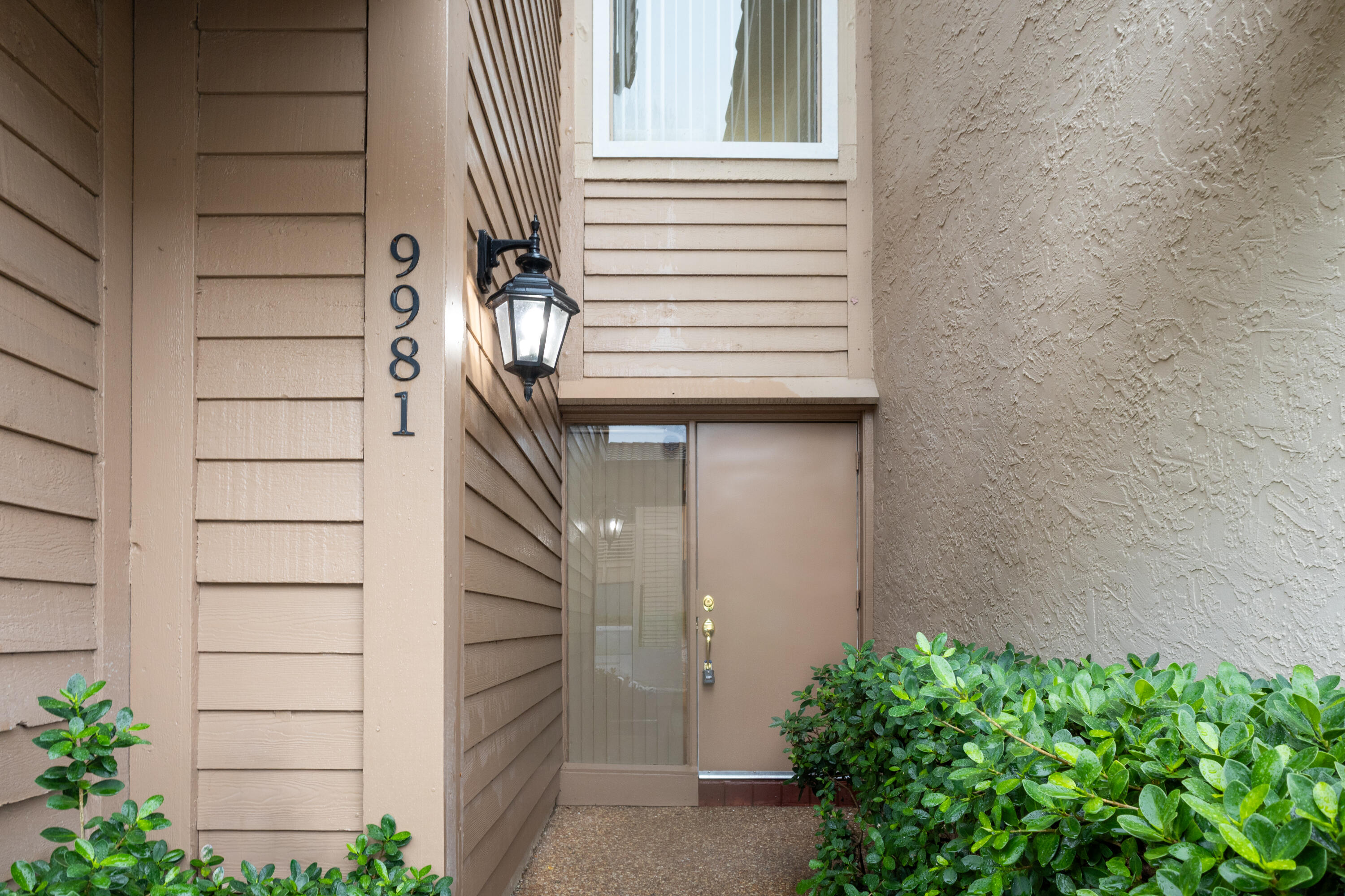 Welcoming entry featuring horizontal siding exterior, illuminated wall sconce, brushed metal door hardware, and lush green landscaping with mature shrubs flanking the entrance Welcoming entry featuring horizontal siding exterior, illuminated wall sconce, brushed metal door hardware, and lush green landscaping with mature shrubs flanking the entrance