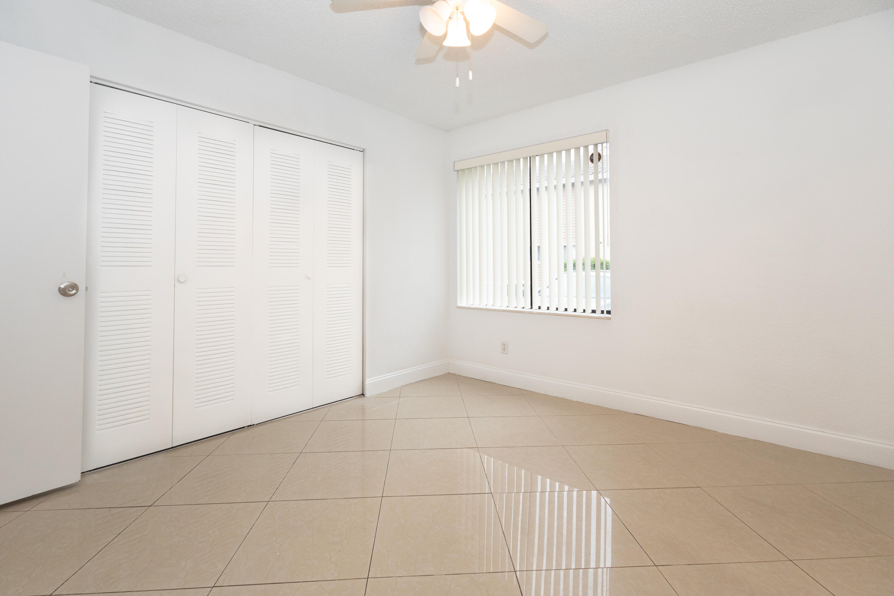Bright and spacious room featuring tile flooring, a ceiling fan with integrated lighting, louvered closet doors, and large windows with vertical blinds allowing ample natural light Bright and spacious room featuring tile flooring, a ceiling fan with integrated lighting, louvered closet doors, and large windows with vertical blinds allowing ample natural light