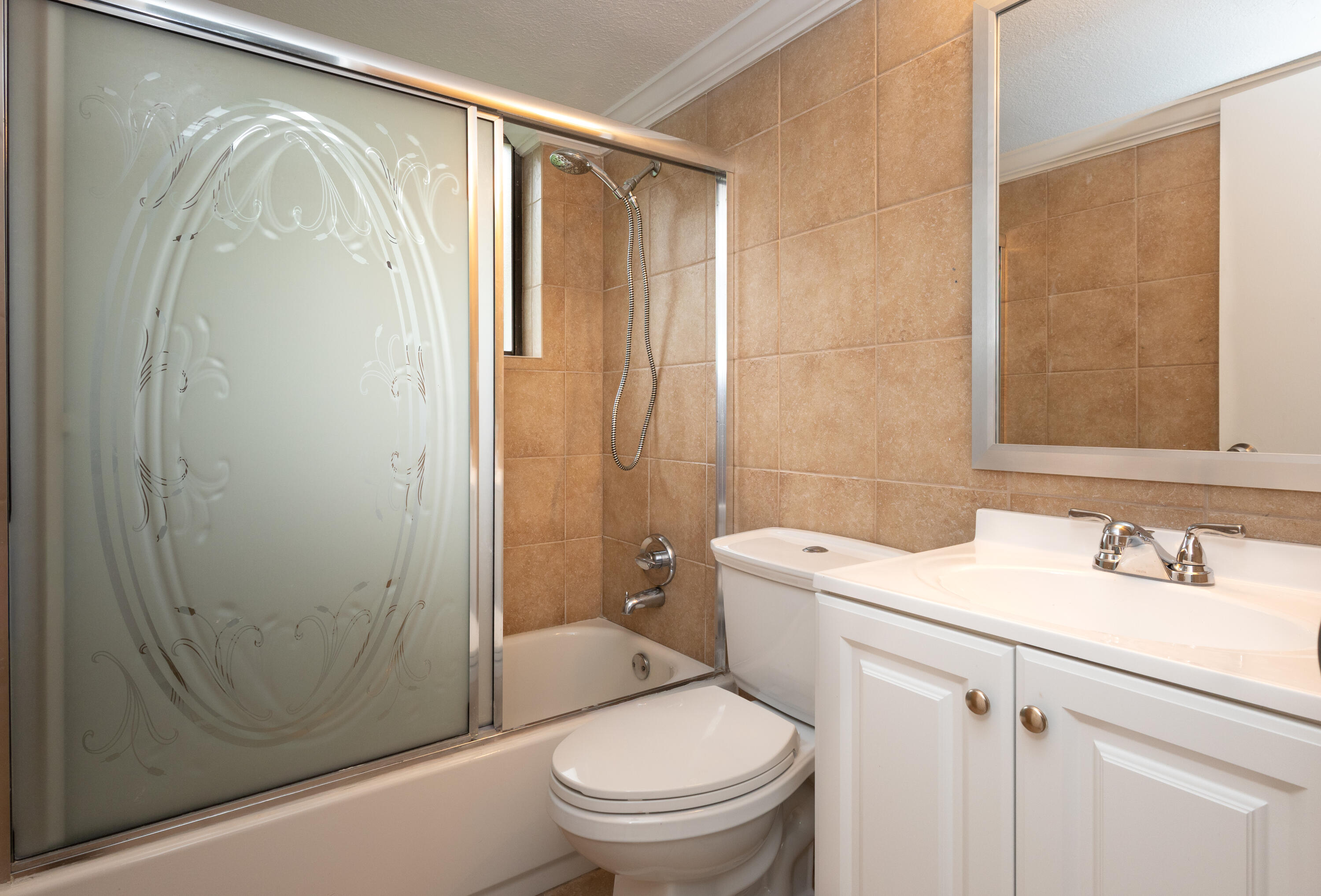 Spa-inspired bathroom featuring warm ceramic tile walls, a frameless glass-enclosed shower with chrome fixtures, white cabinetry vanity with dual-sink configuration, and a white porcelain toilet