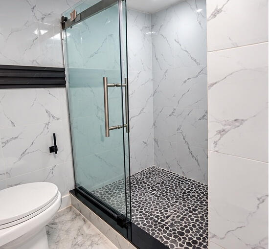 Walk-in shower featuring a frameless glass enclosure, pebble-stone flooring, large-format veined tile surround, and matte black hardware