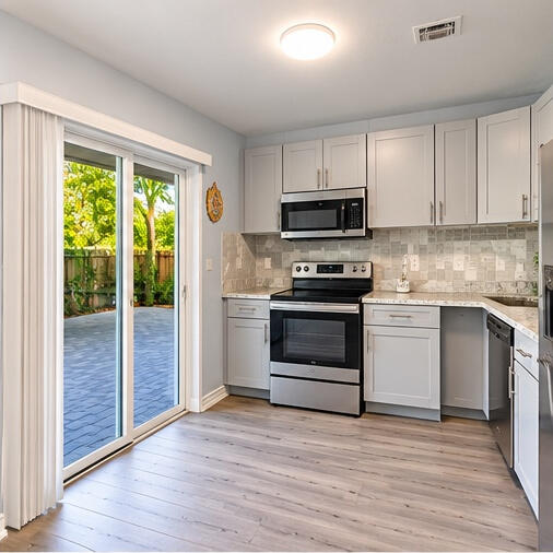 Kitchen featuring shaker-style cabinetry, stainless steel appliances, stone-look countertops, patterned tile backsplash, and wood-finish flooring with sliding glass door access to a paved patio