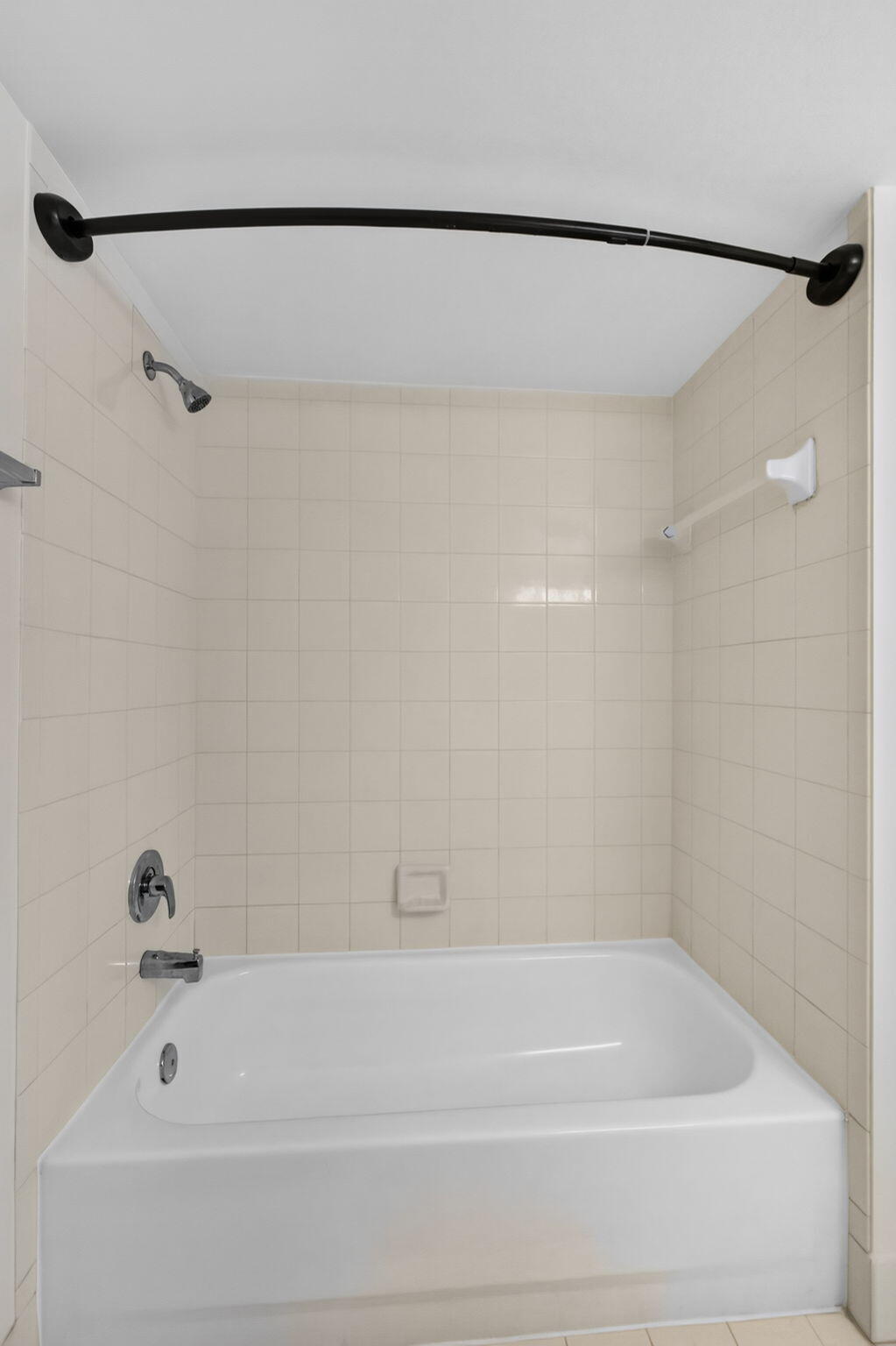 Bathtub with a white acrylic surround, ceramic wall tiling, polished chrome faucet, and a matte black curved shower rod