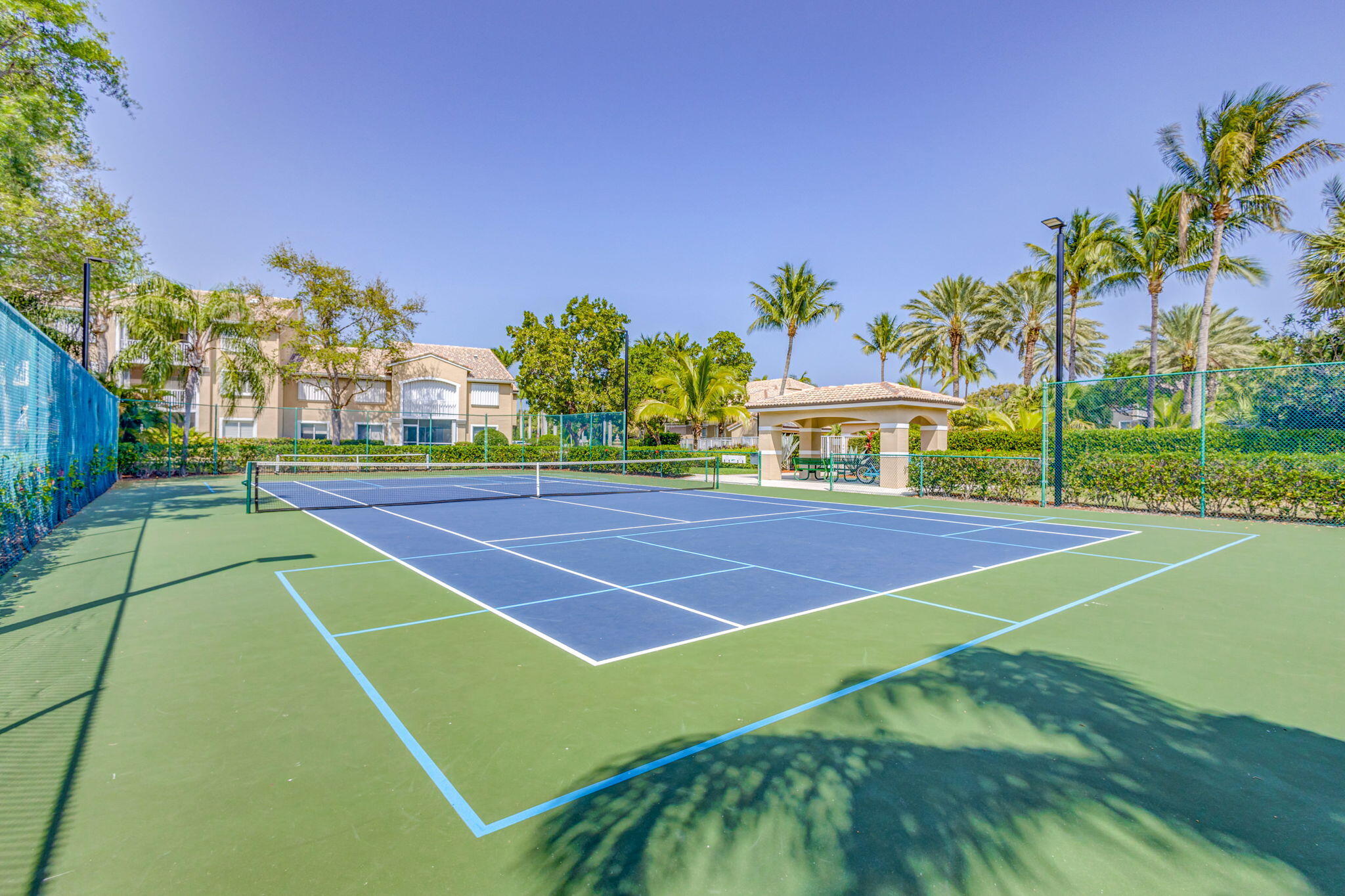 Hard-surfaced tennis court featuring perimeter chain-link fencing, dual-tone court markings, and an integrated pavilion with a tiled roof