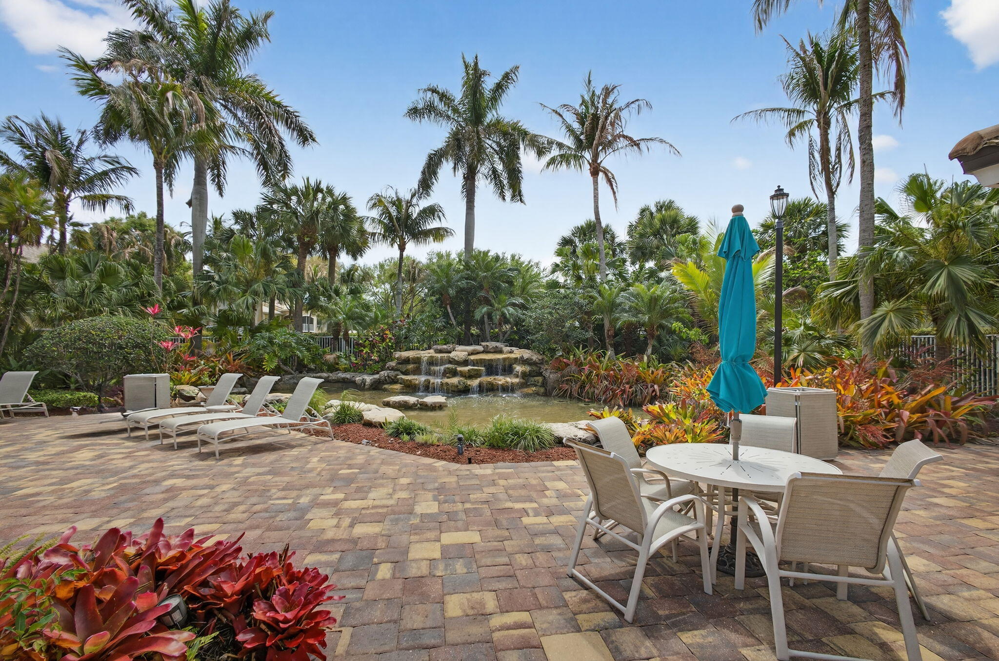 Paver patio featuring a multi-tier rock waterfall, mature palm trees, tropical landscaping, and integrated landscape lighting