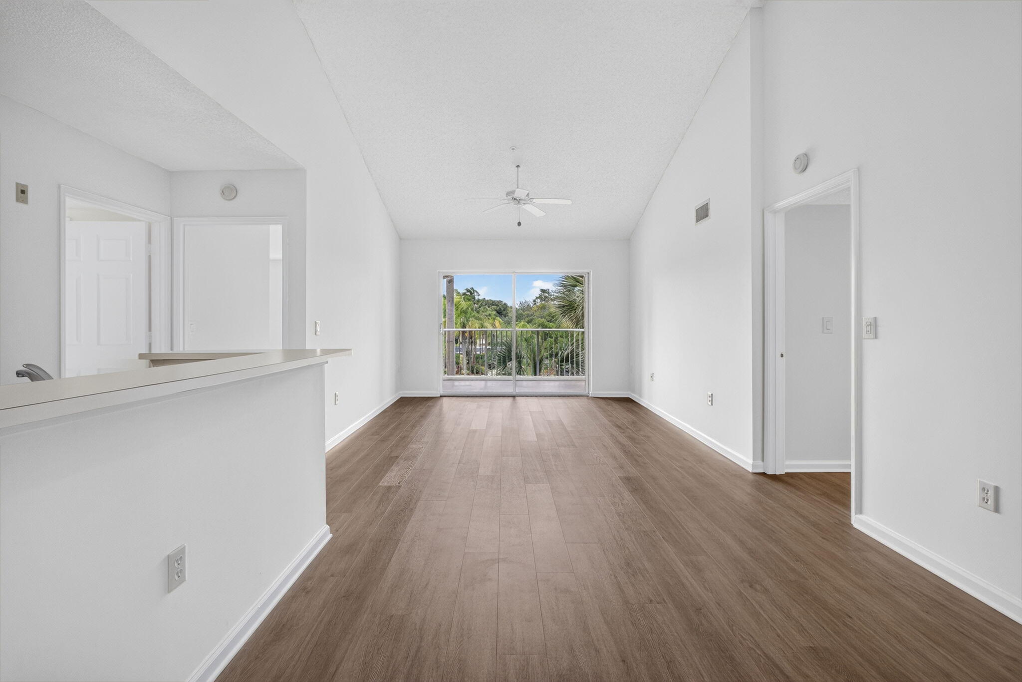 Vaulted ceilings featuring a central ceiling fan, vinyl plank flooring, a pass-through bar counter, and sliding glass doors with balcony access.