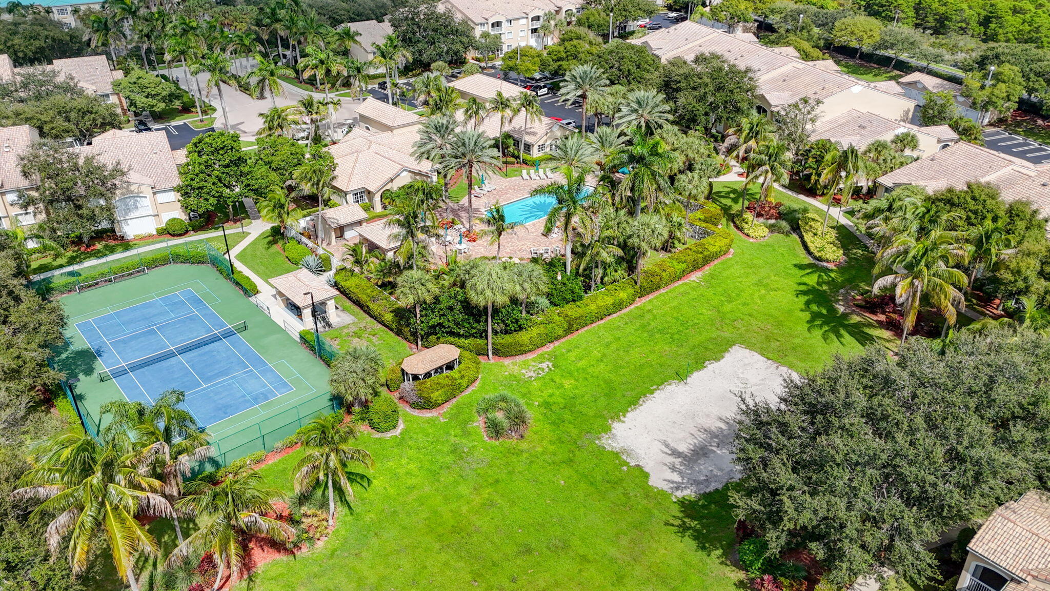 Aerial perspective of community amenities featuring a fenced tennis court, swimming pool, manicured lawn, mature palm trees, and covered pavilion